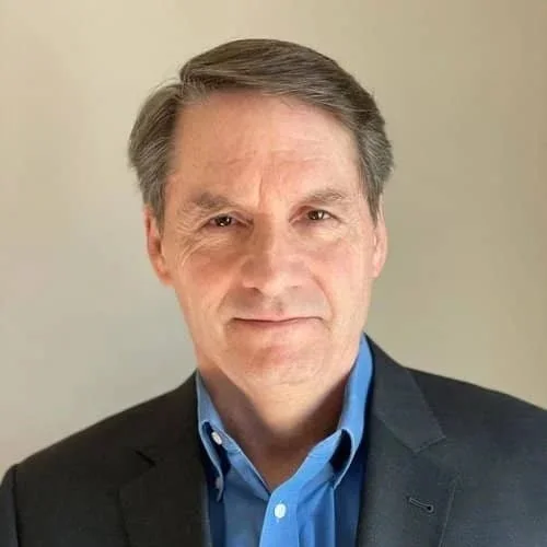 A middle-aged man with short gray hair wearing a black blazer and blue dress shirt standing against a plain light-colored wall.