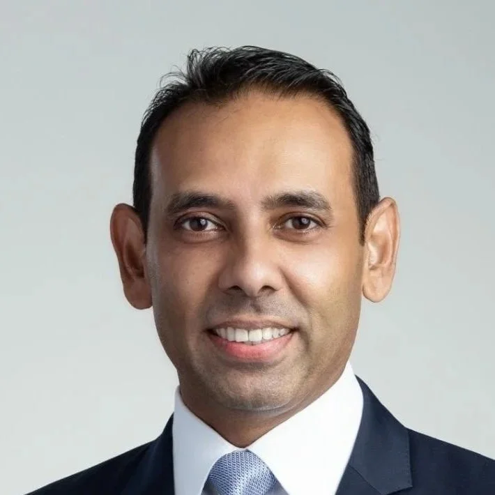 Professional headshot of a man in a dark suit, white shirt, and light blue tie, smiling against a plain light background.
