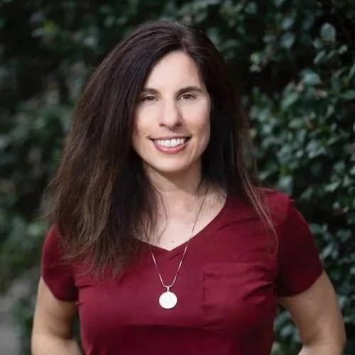 A woman with long brown hair wearing a maroon shirt and a silver necklace, standing outdoors in front of green foliage, smiling at the camera.