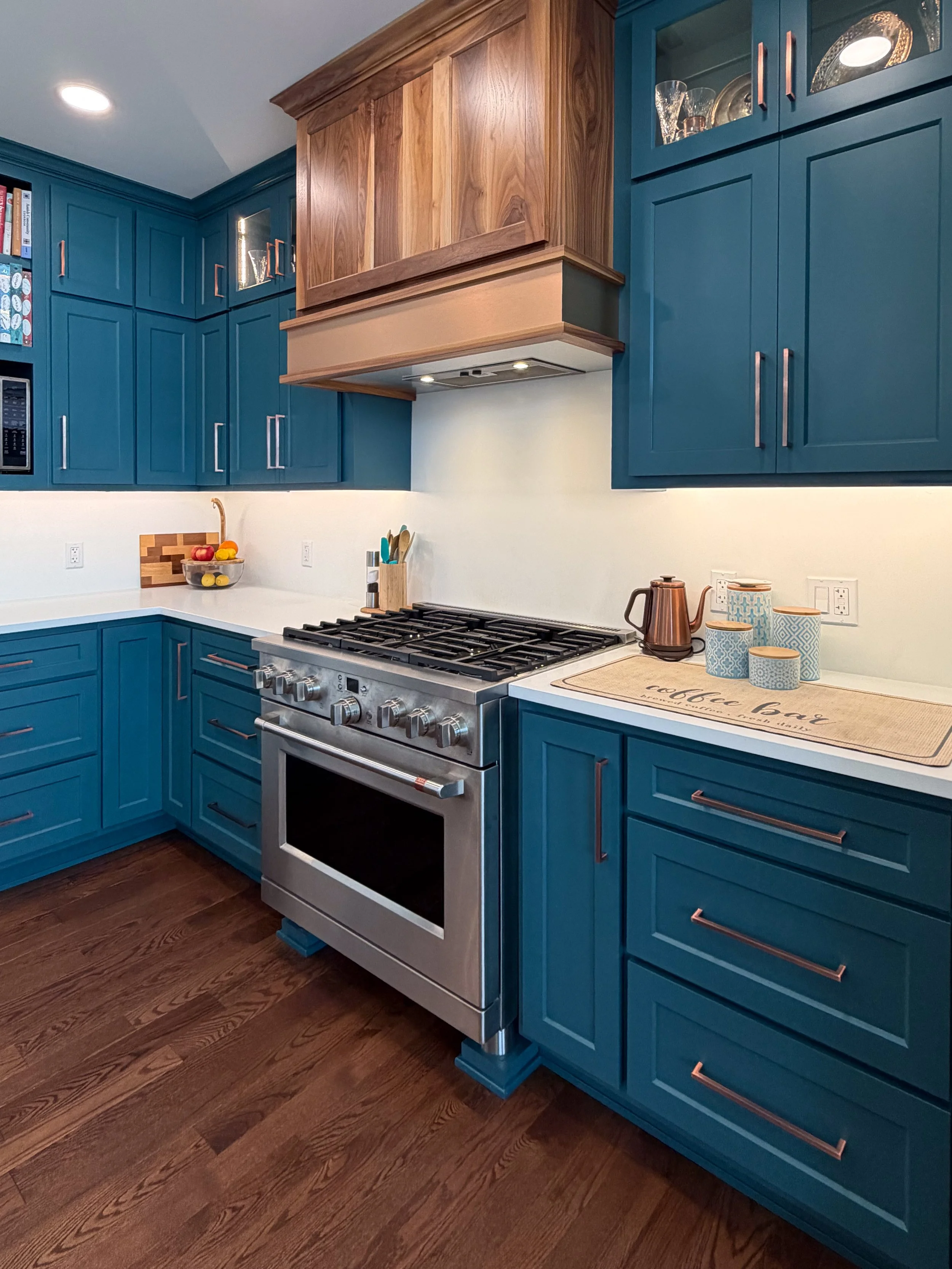 Modern kitchen featuring deep teal shaker-style cabinets with copper handles, a professional stainless steel gas range, and a custom natural wood range hood.
