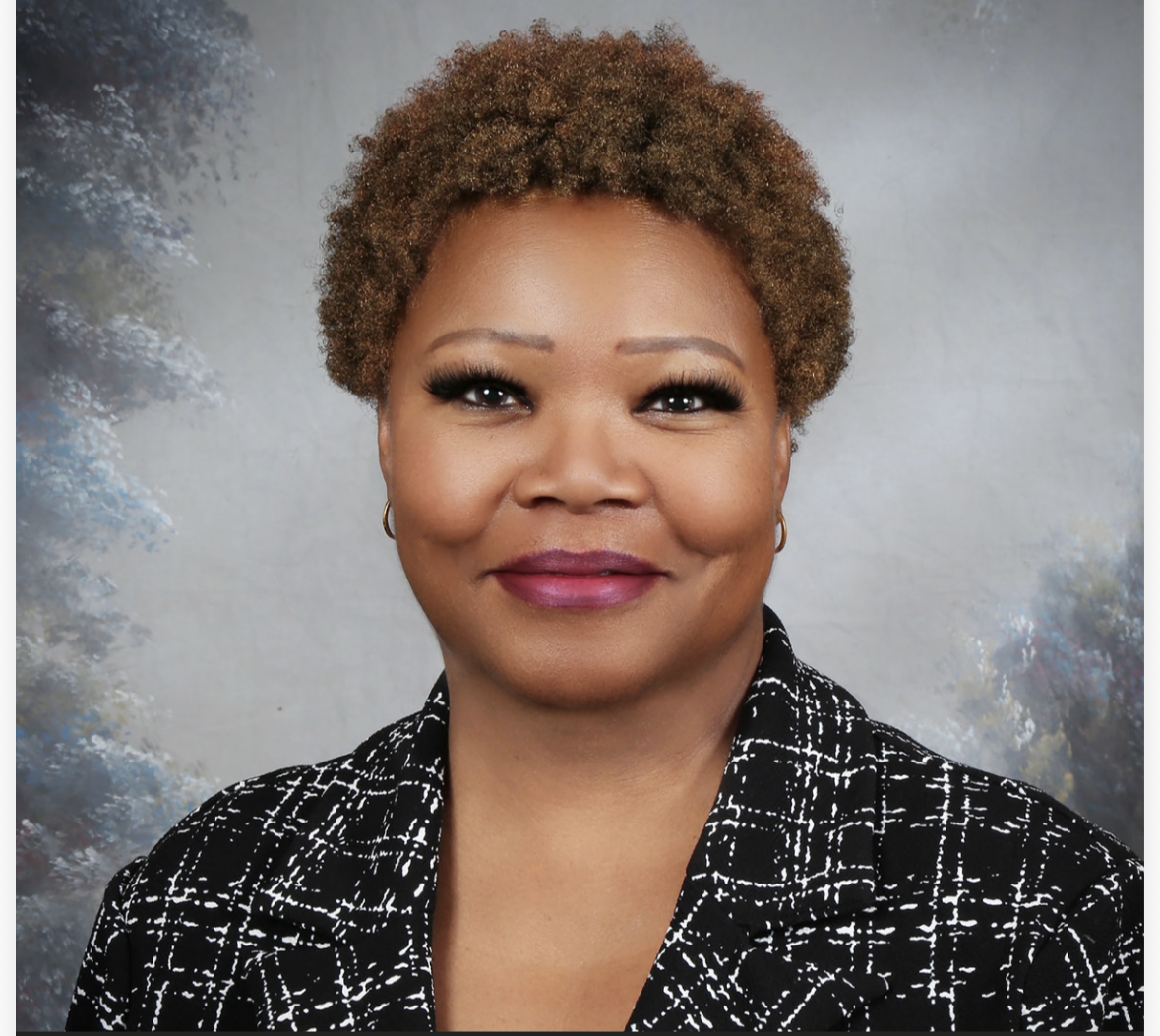 A woman with short curly hair smiling at the camera, wearing a black and white patterned top and gold hoop earrings, against a soft cloudy background.