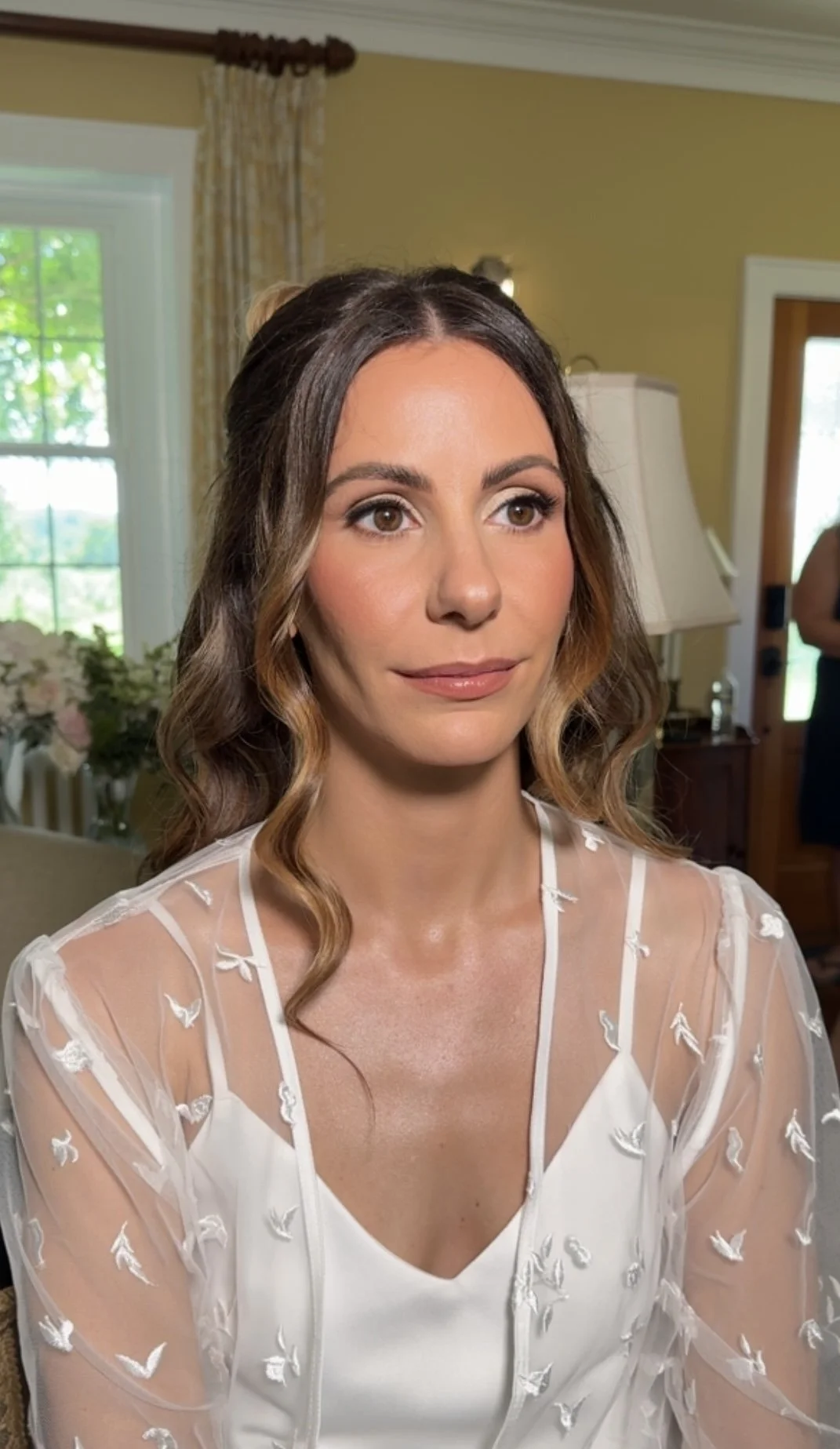 A woman with wavy brown hair and light makeup, wearing a white dress with sheer fabric and embroidered birds, in a well-lit room with yellow walls, a window, and a lamp.