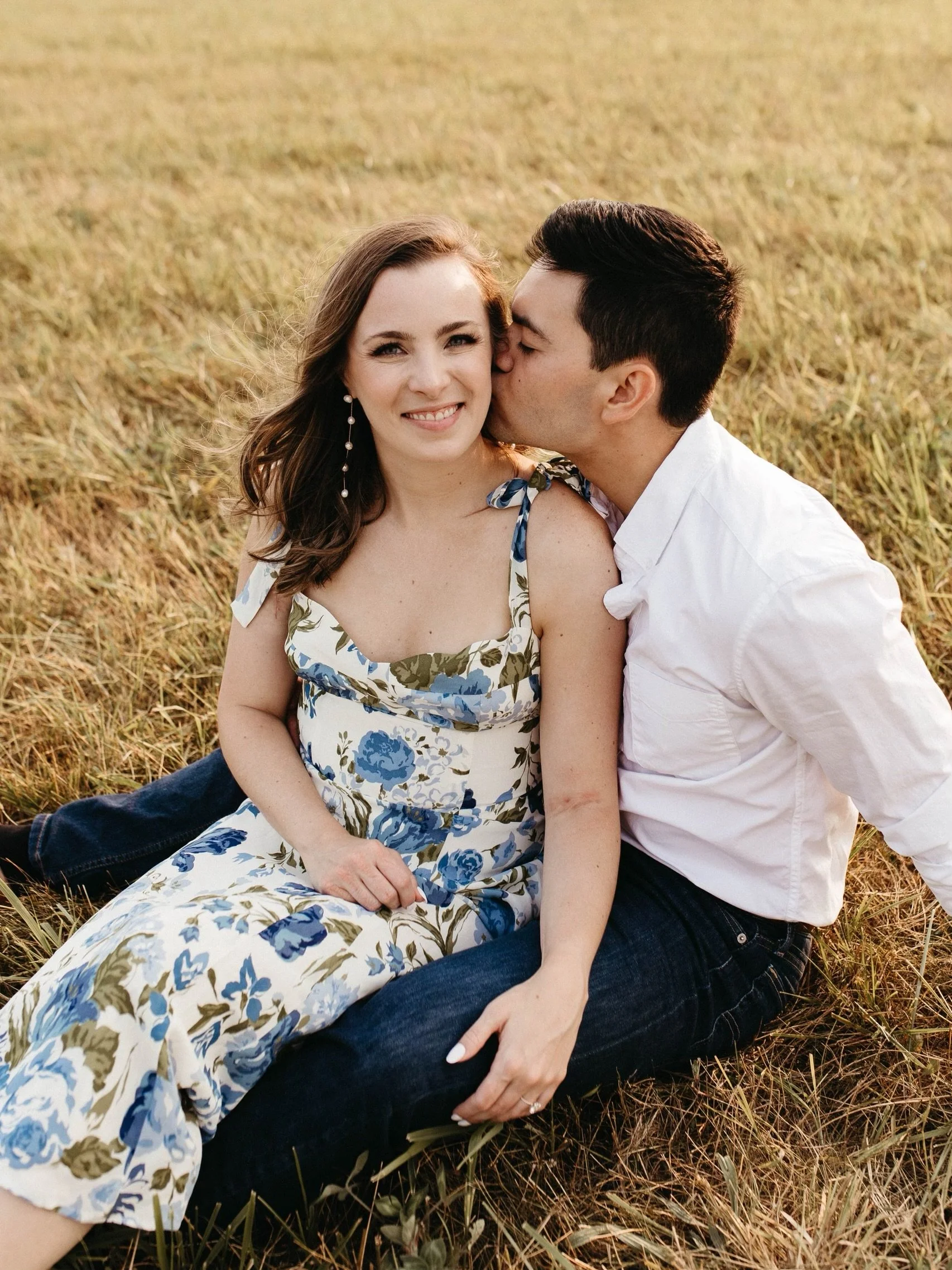 A couple sitting on the grass, the man is kissing the woman's cheek, both smiling and dressed casually.