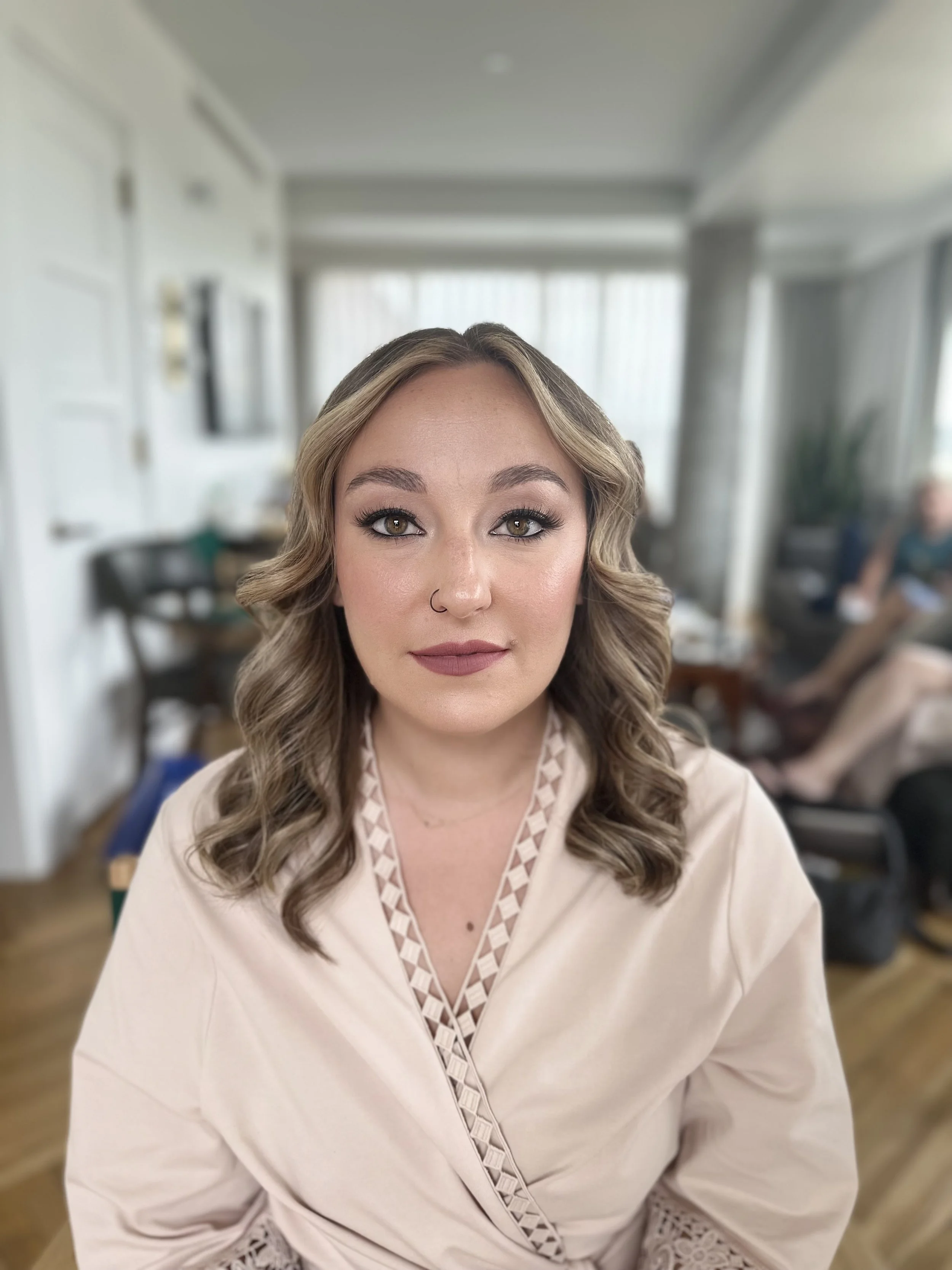 A woman with light brown, wavy hair and light brown eyes, wearing makeup and a nose ring, sitting indoors with a blurred background of a room with large windows and several people.
