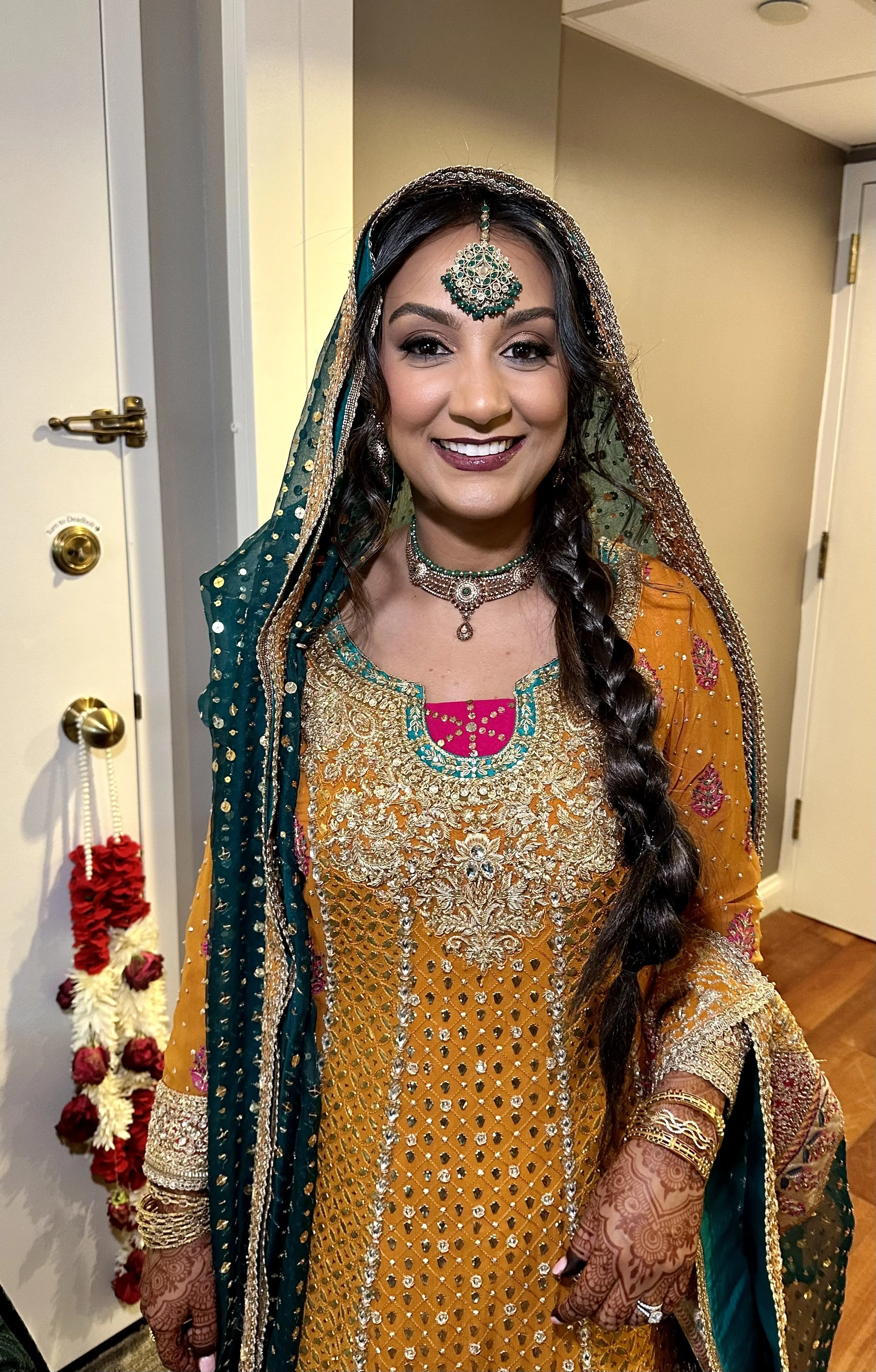 Woman in traditional Indian attire, including embroidered dress, dupatta with embellishments, jewelry, and henna on her hands, smiling at the camera.
