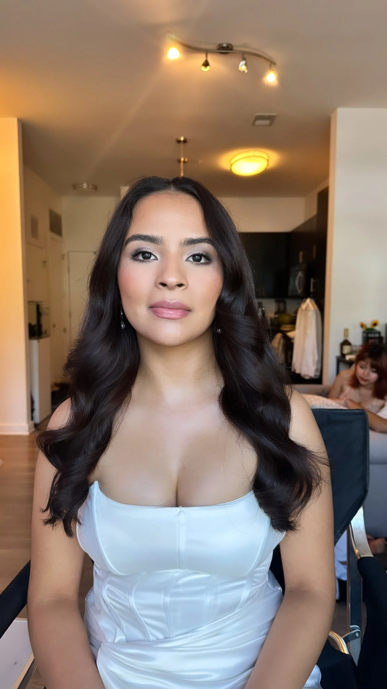 A woman with long, wavy dark hair and makeup, wearing a white satin dress, sitting indoors in a well-lit room with a person in the background.