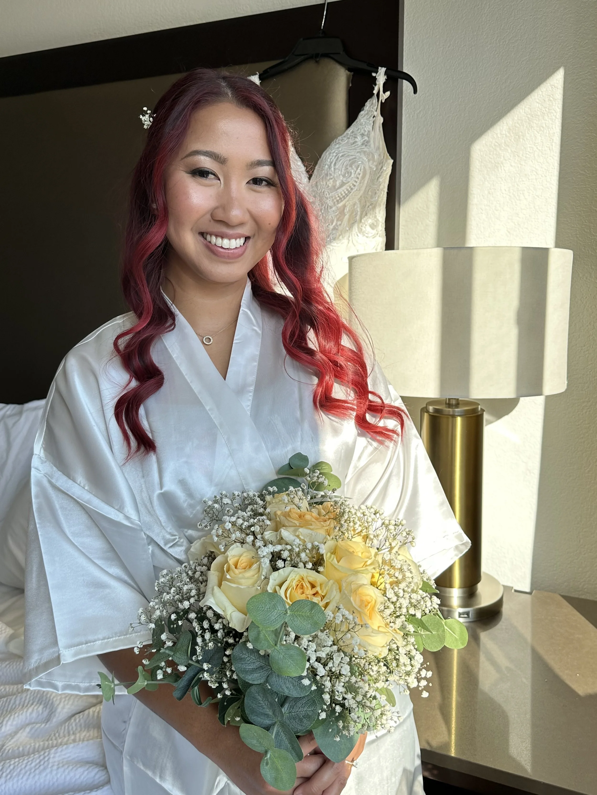 A woman with long red hair smiling while holding a bouquet of yellow roses and white baby's breath. She is wearing a white satin robe and standing near a beige lamp, with a wedding dress hanging behind her on a hanger.