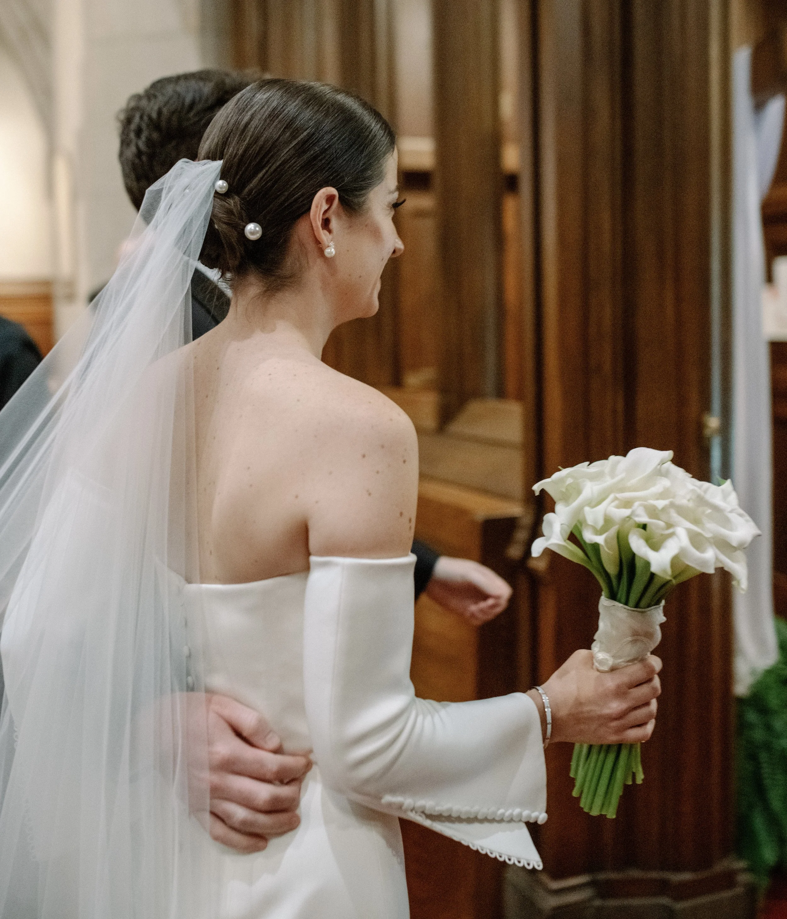 A bride in a white off-the-shoulder wedding dress holding a bouquet of white calla lilies during a wedding ceremony inside a wooden church, with a man embracing her from behind.