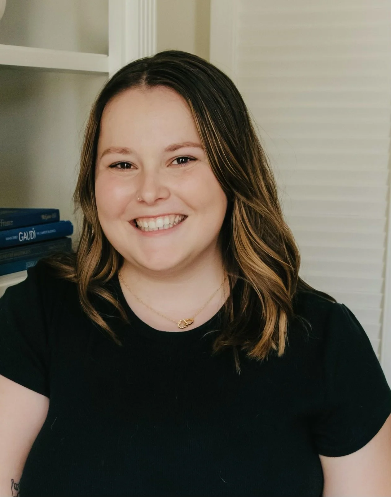 A smiling woman with shoulder-length brown hair with highlights, wearing a gold necklace with a pendant, and a black shirt, standing indoors near a bookshelf.