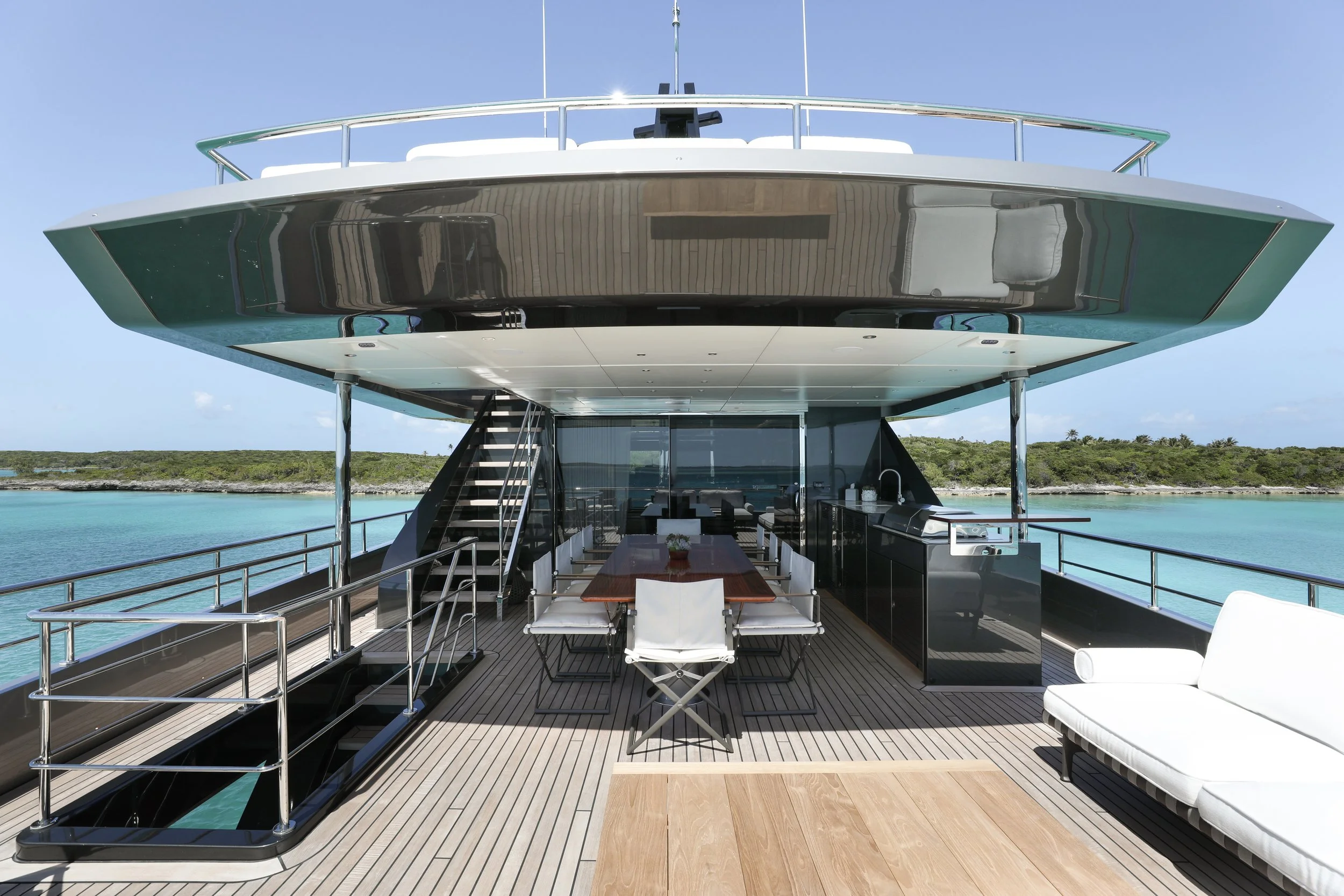 Luxury yacht with a dining table and chairs on deck, surrounded by turquoise water and green shoreline under a blue sky.