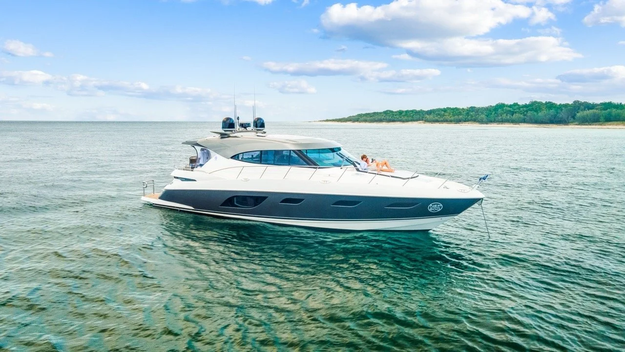 A white yacht floating on calm water near a distant green shoreline with trees under a partly cloudy sky.