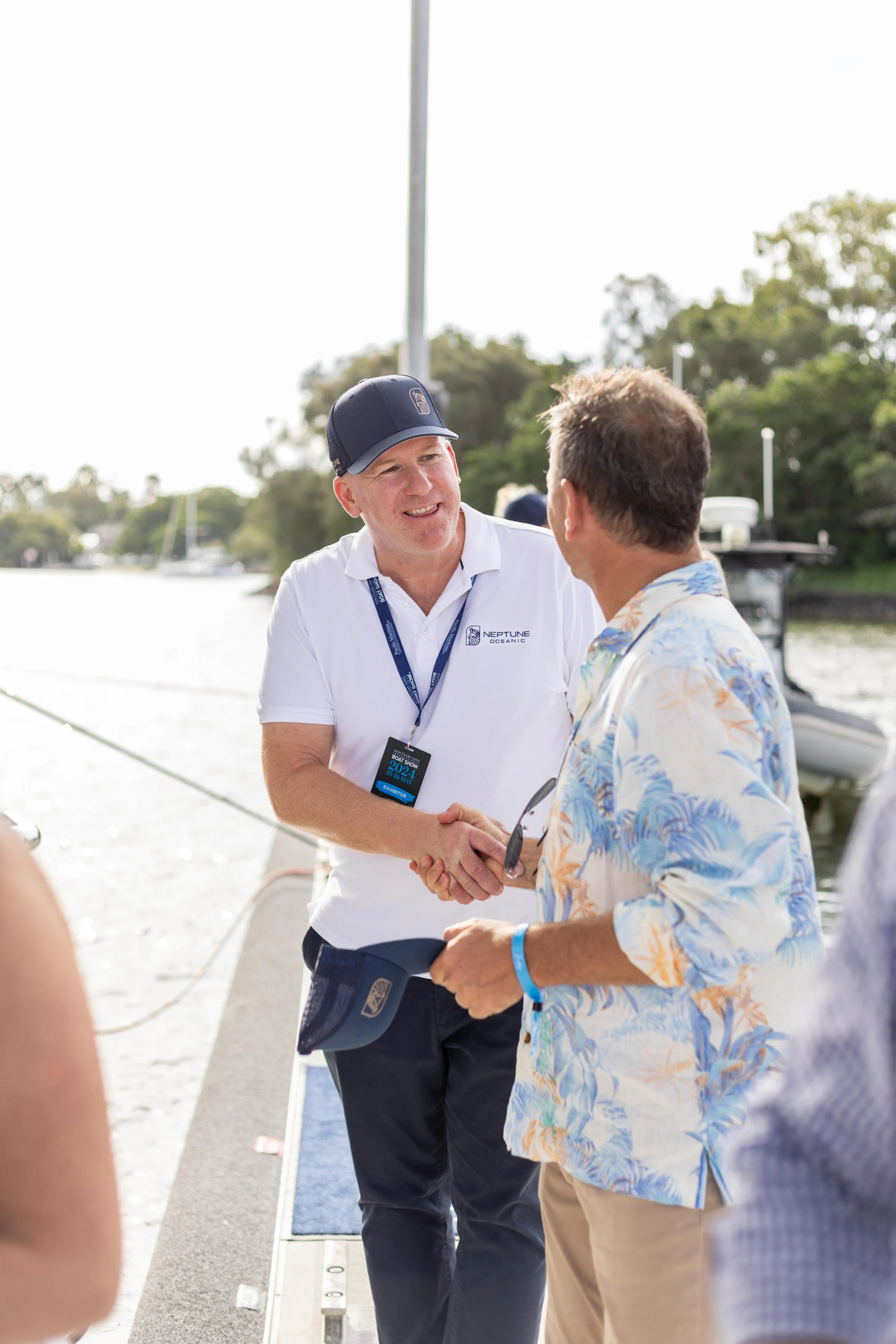 Two men shaking hands near a boat dock, with a marina and sailboats in the background, during daytime.
