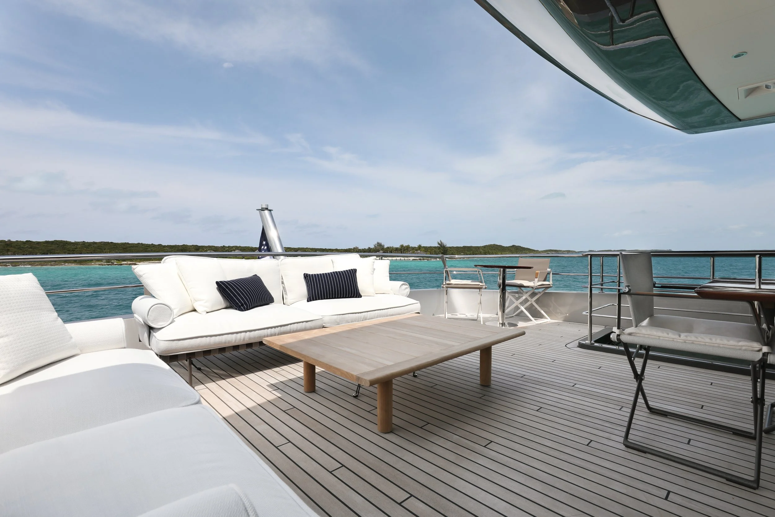 Outdoor deck area with white cushioned sofas, a wooden table, chairs, and a view of water and land in the distance, under a blue sky with some clouds.