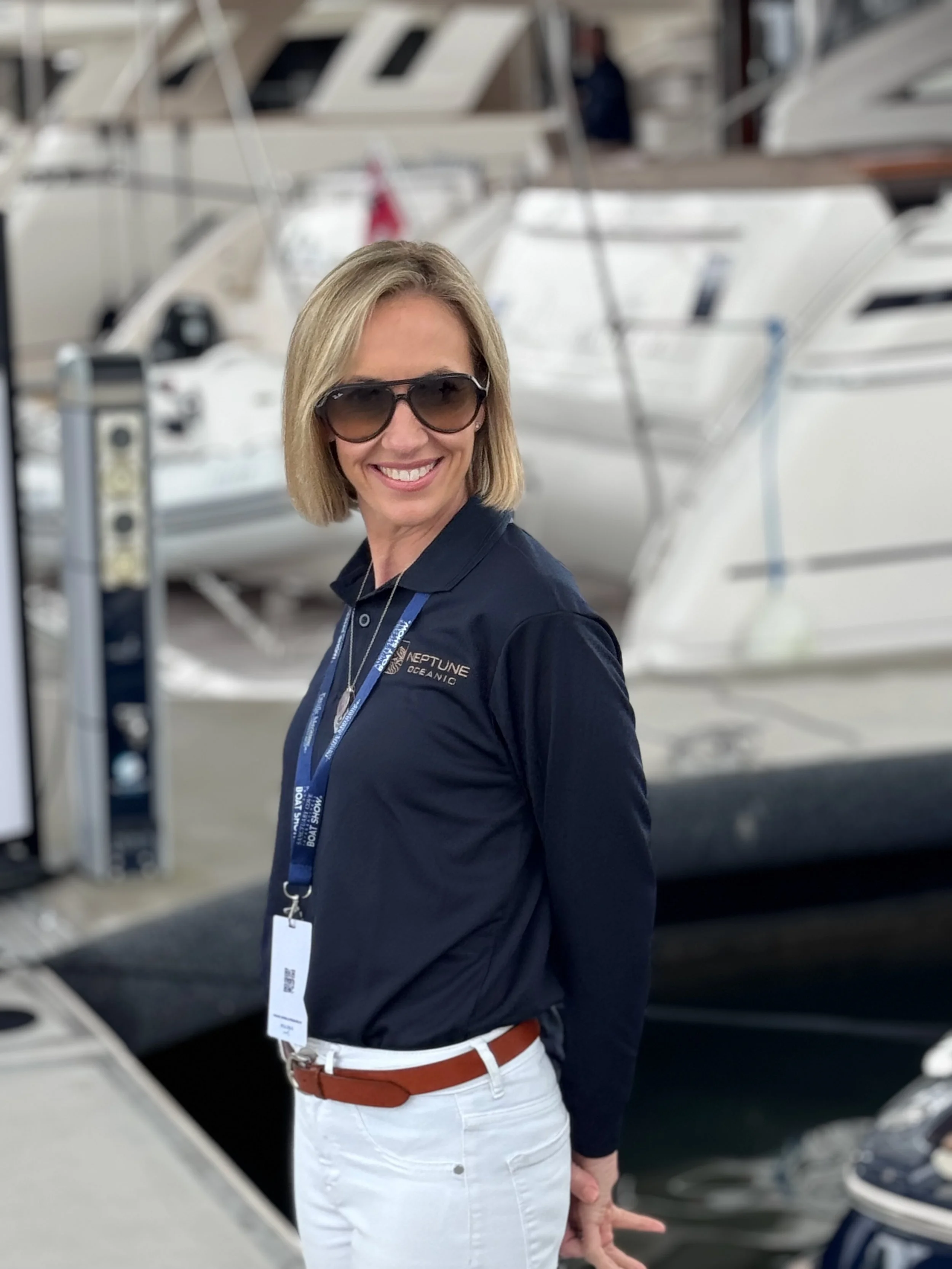 A woman smiling and wearing sunglasses at a marina, surrounded by white yachts and boats.