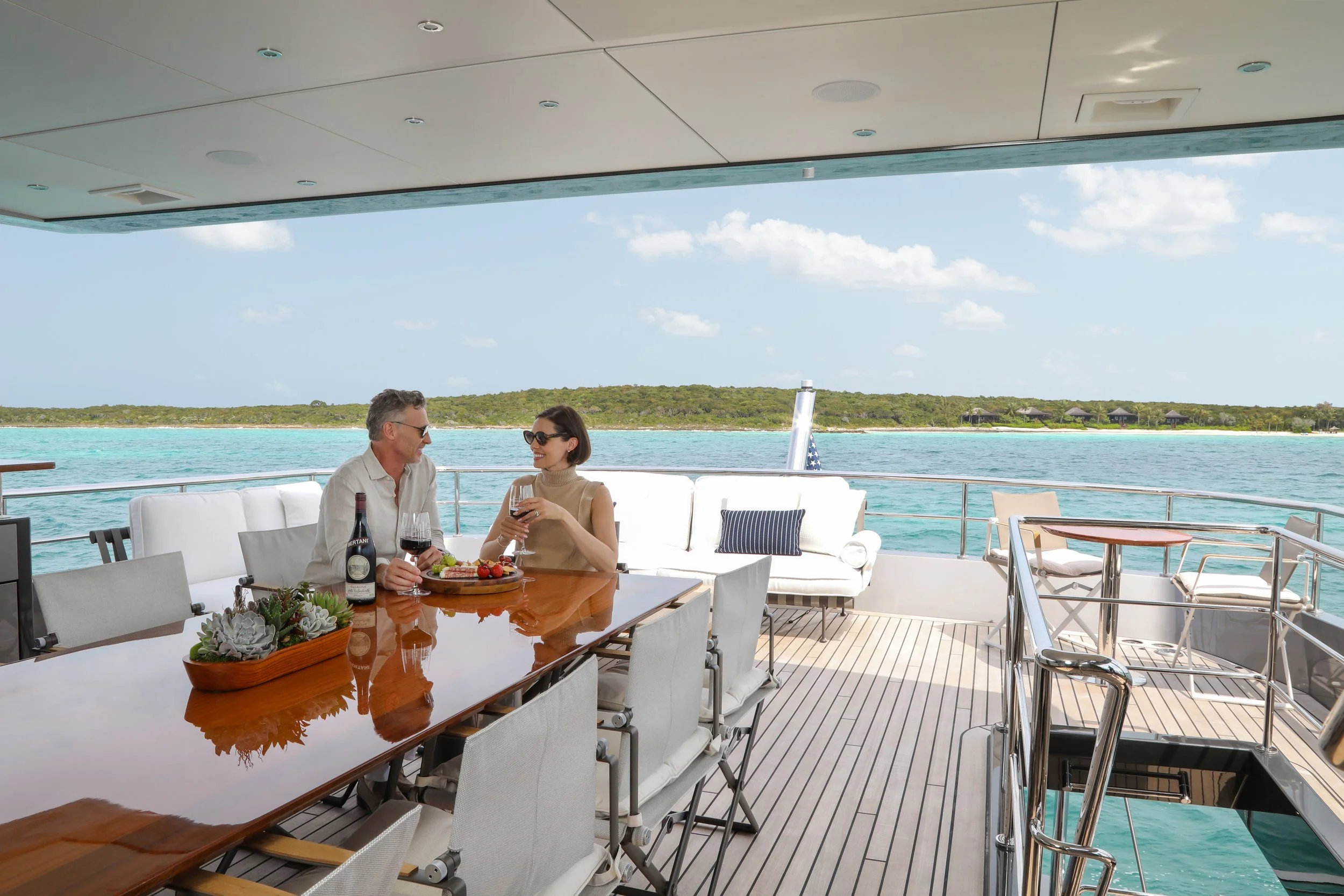 Two people enjoying a meal and wine on the deck of a yacht with ocean and island in the background.