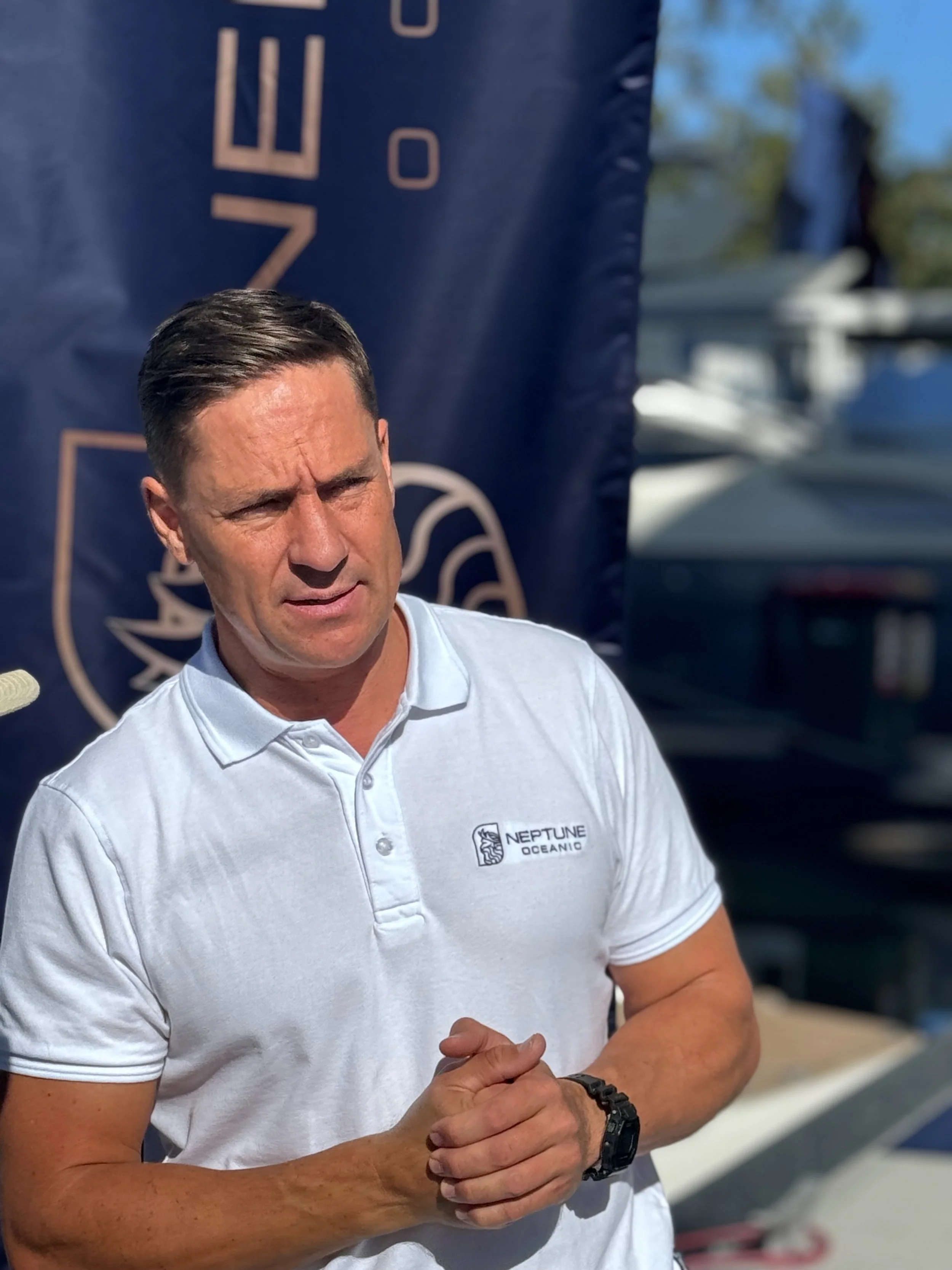 A man with dark hair wearing a white polo shirt with 'Neptune Oceanic' logo standing outdoors, with boats in the background.