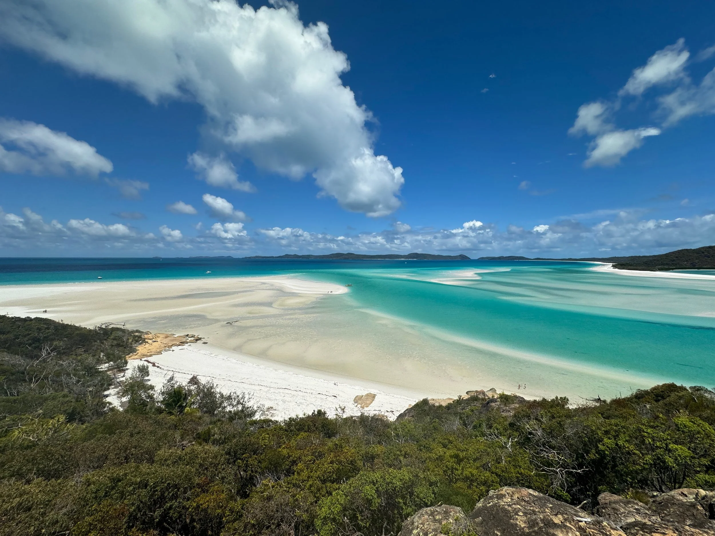 Scenic view of a beach with clear turquoise waters, white sand, lush green vegetation in the foreground, and a partly cloudy blue sky.