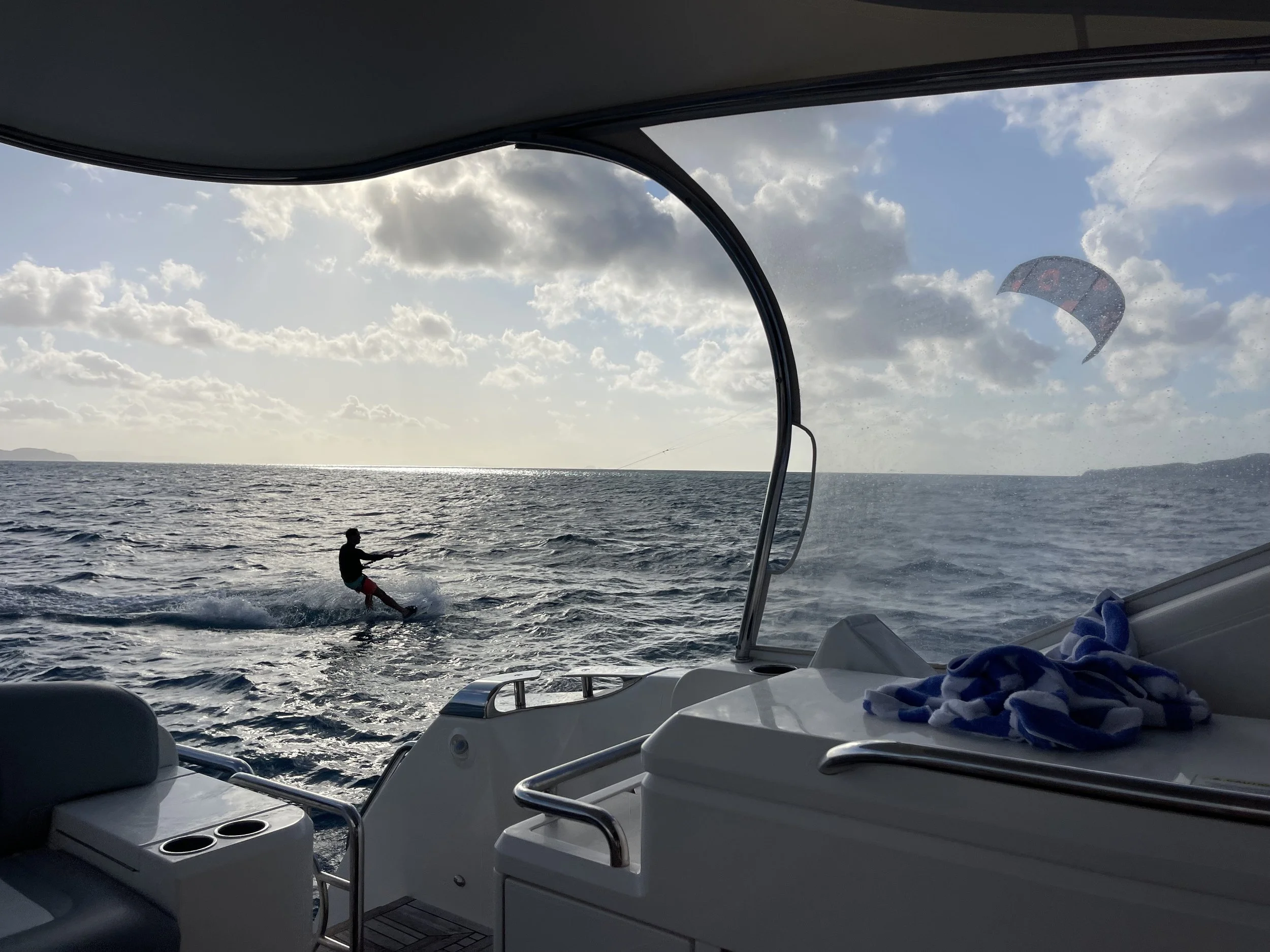 View from a boat showing a person wakeboarding on the water with a kite in the sky, during a partly cloudy day.
