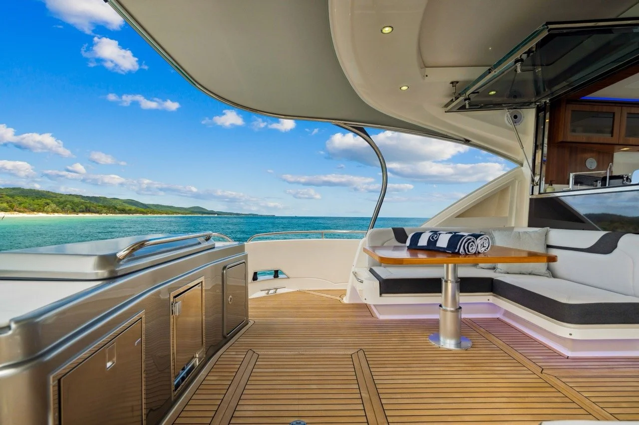 The deck of a yacht with outdoor seating, a small table with rolled towels, and a view of the ocean and distant shoreline under a blue sky with clouds.