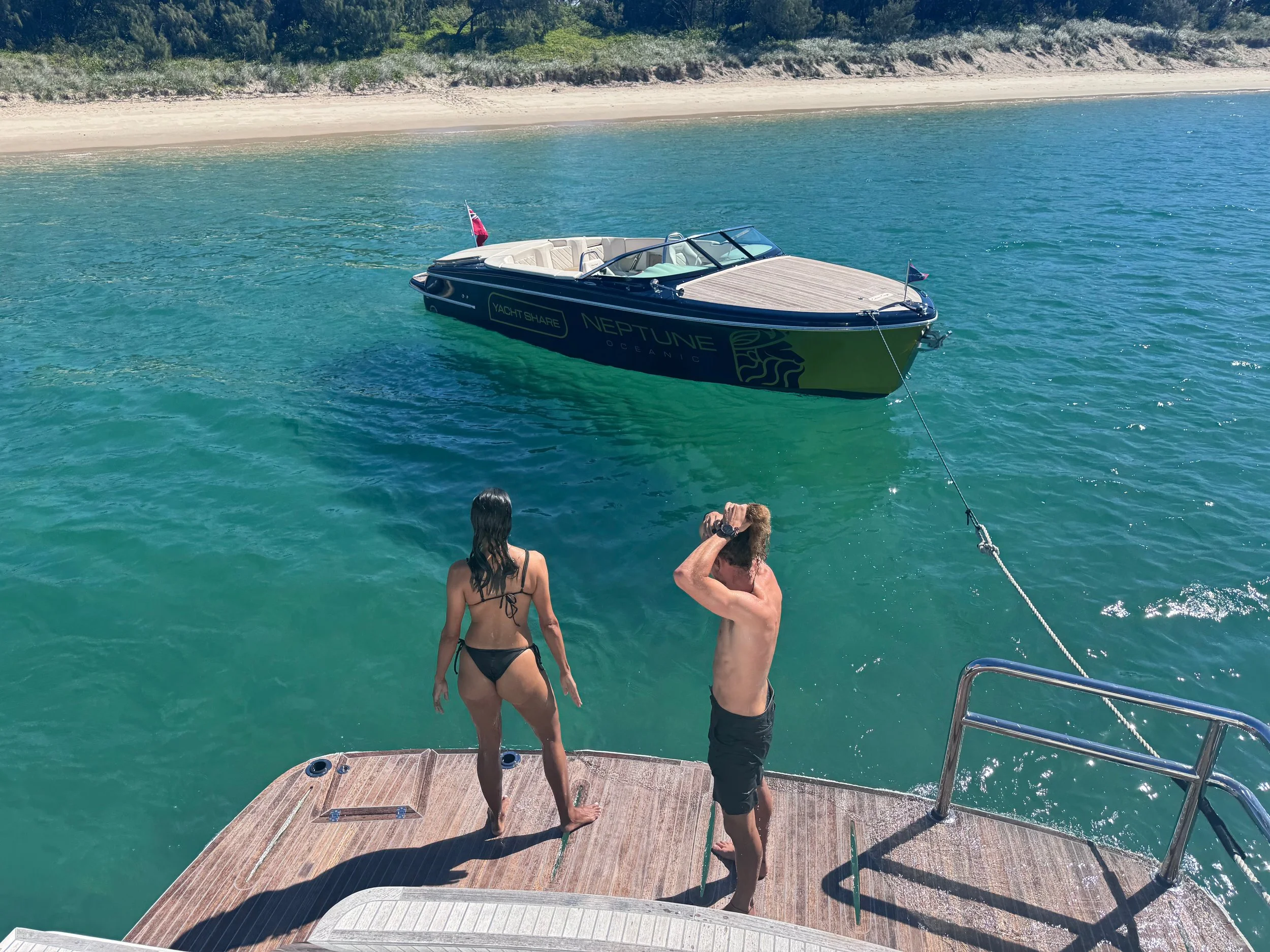Two people standing on the deck of a boat looking at a yacht anchored near a sandy beach with trees in the background.
