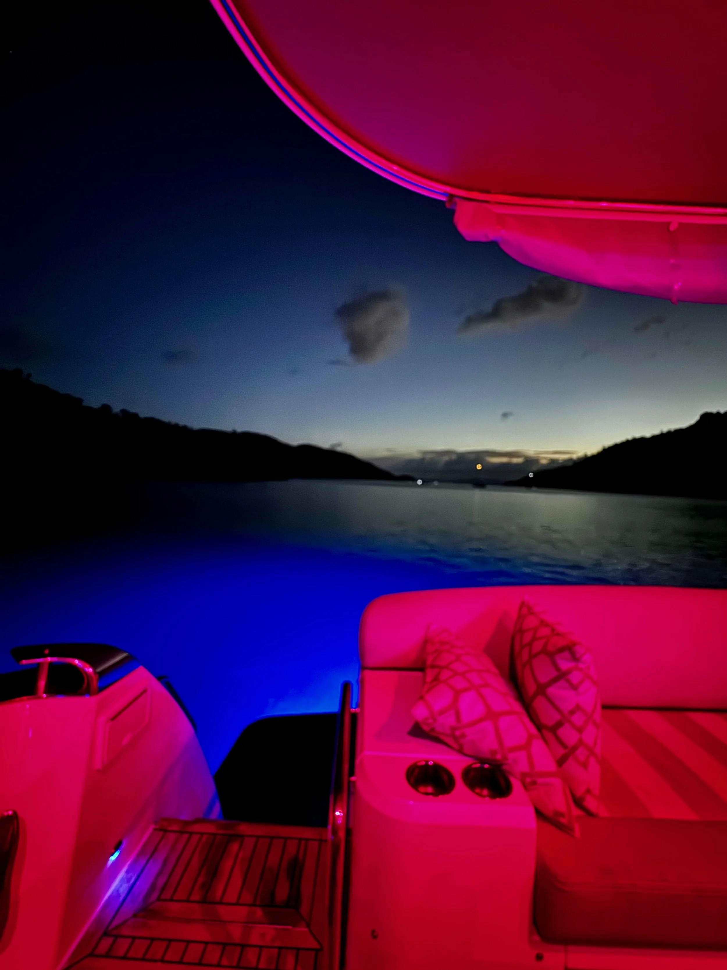Nighttime scene on a boat with pink and blue LED lights, overlooking a calm river or lake with silhouetted hills and few clouds in the sky.