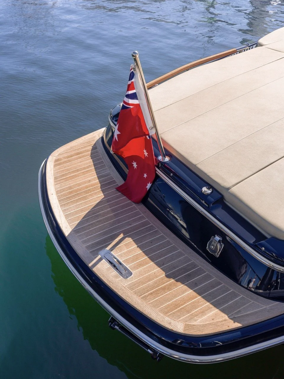 Close-up of the stern of a boat with a wooden deck, a beige cushion, and an Australian flag flying at the back, floating on calm water.