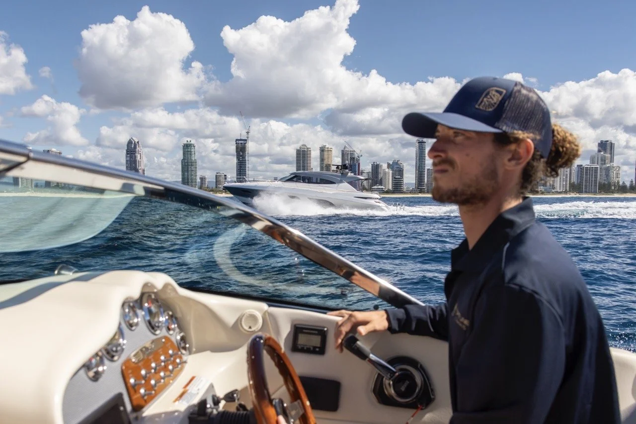 Man steering a boat on the water with modern city skyline and another boat in the background on a sunny day.