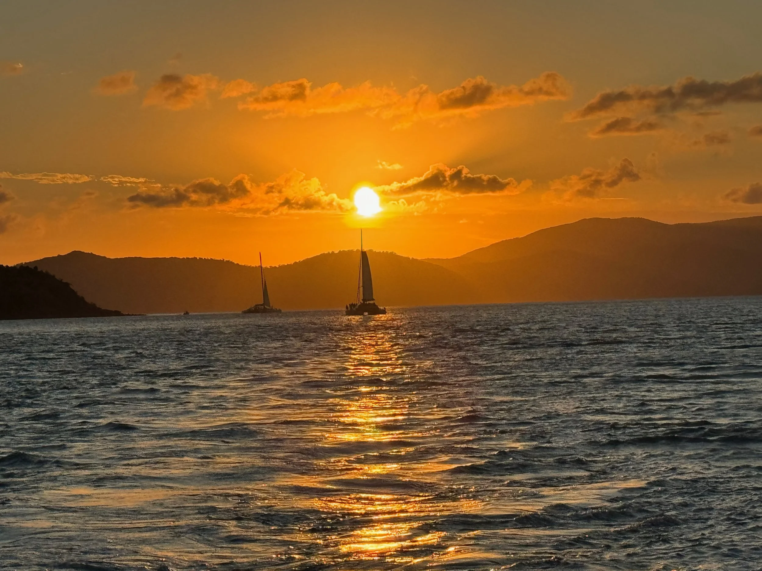 A sunset over a body of water with two sailboats and mountains in the background.