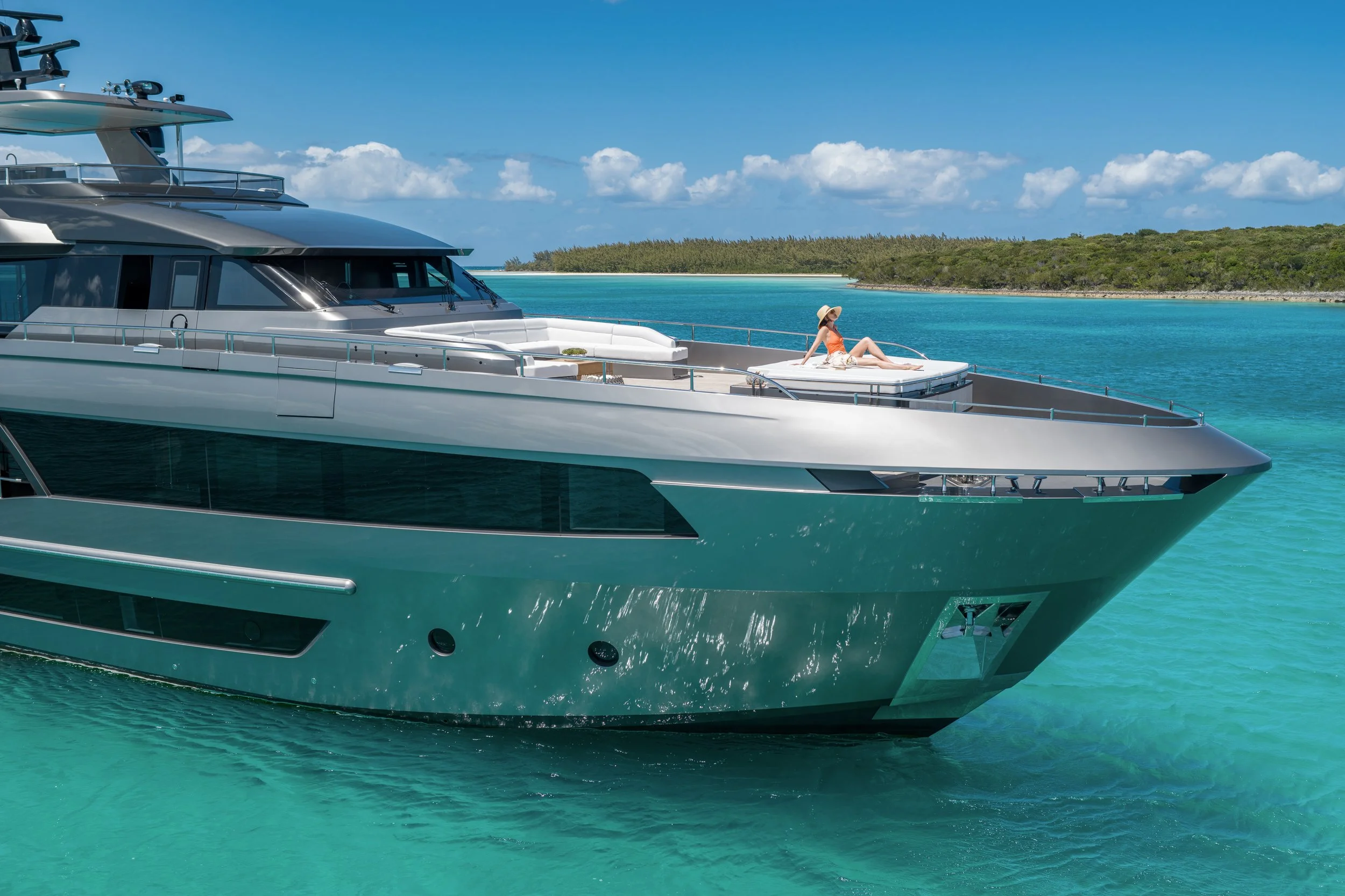 A large luxury yacht floating on turquoise water with a woman relaxing on the sun deck, wearing a hat and orange swimsuit, with a green island and blue sky with clouds in the background.
