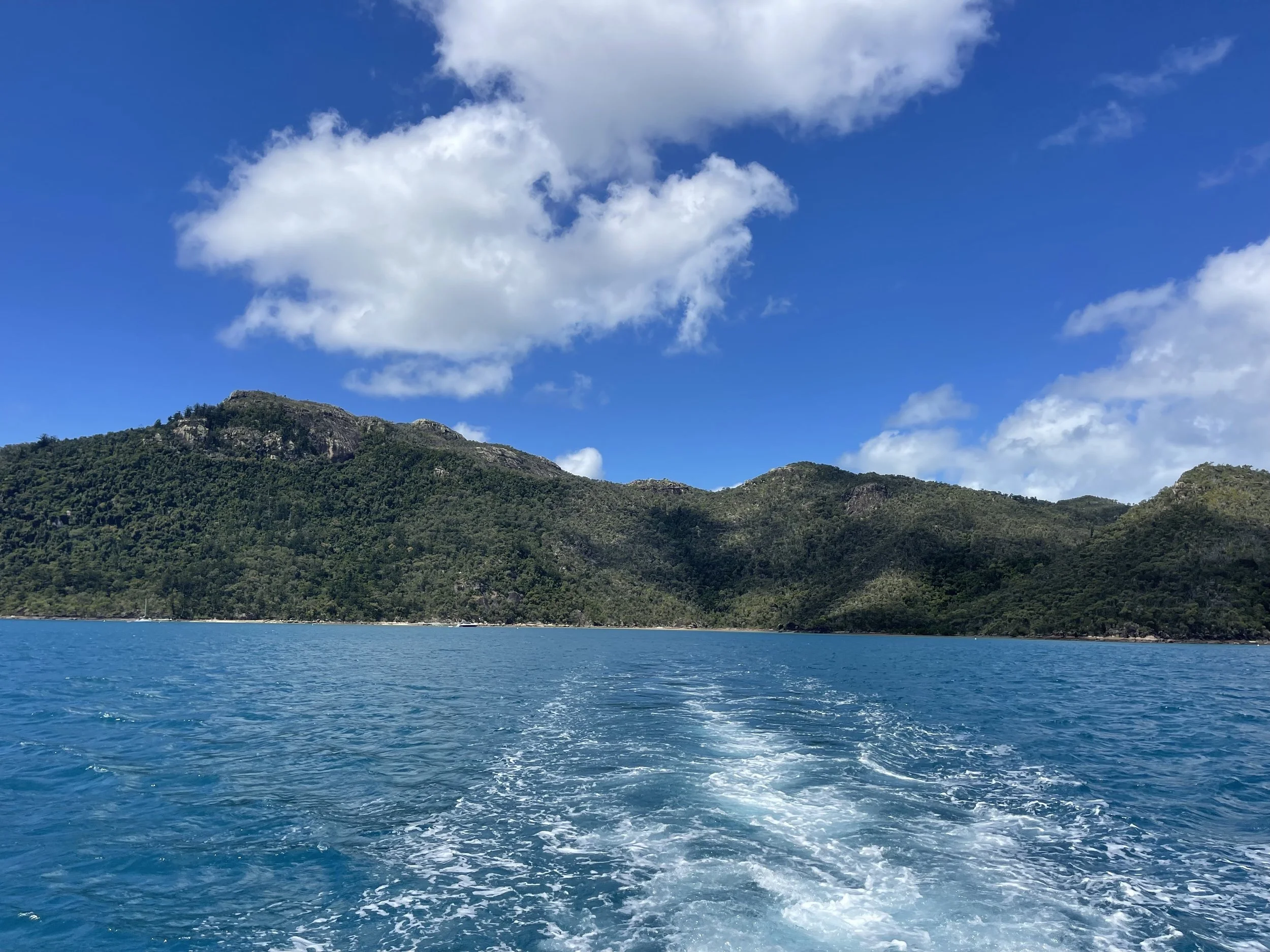 View of a lush green mountain range over calm blue ocean waters with a partly cloudy blue sky.