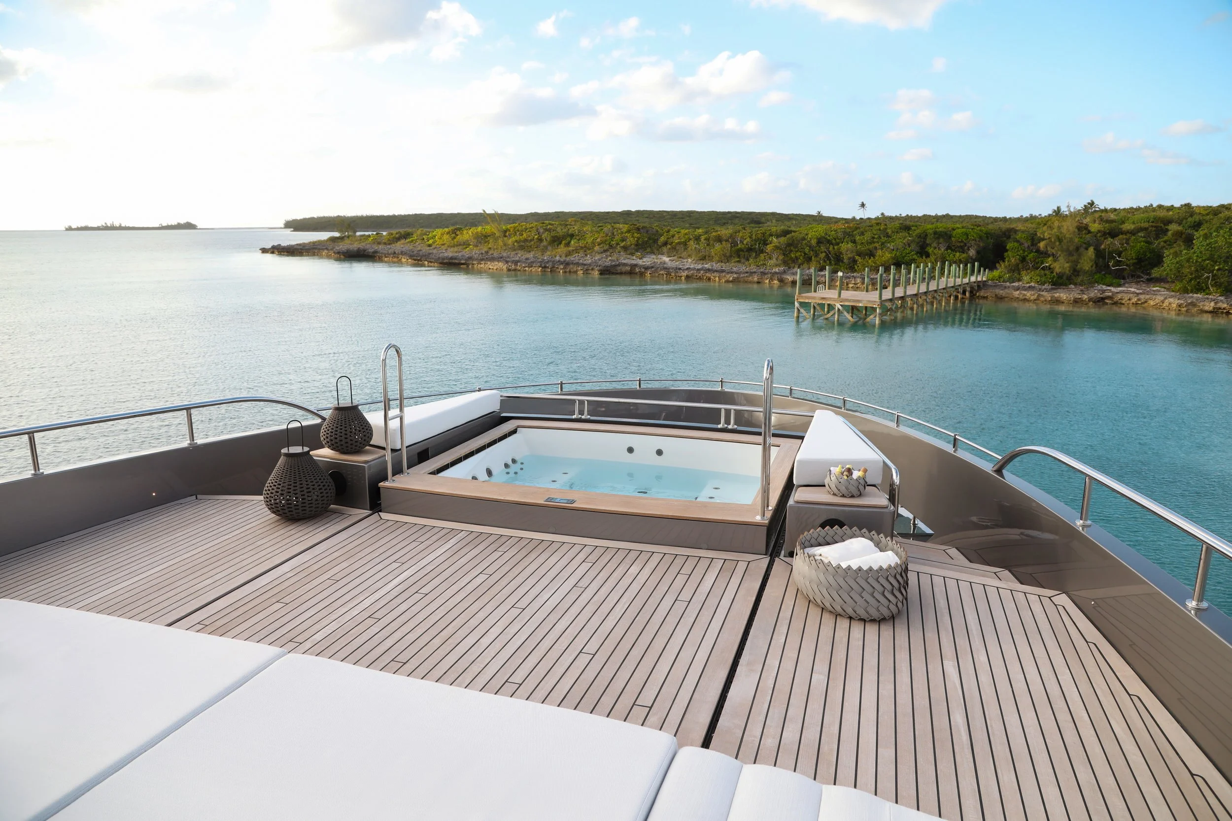 Yacht deck with a hot tub, white cushions, black lanterns, and a basket of towels, overlooking a calm body of water with a dock and green shoreline under a partly cloudy sky.