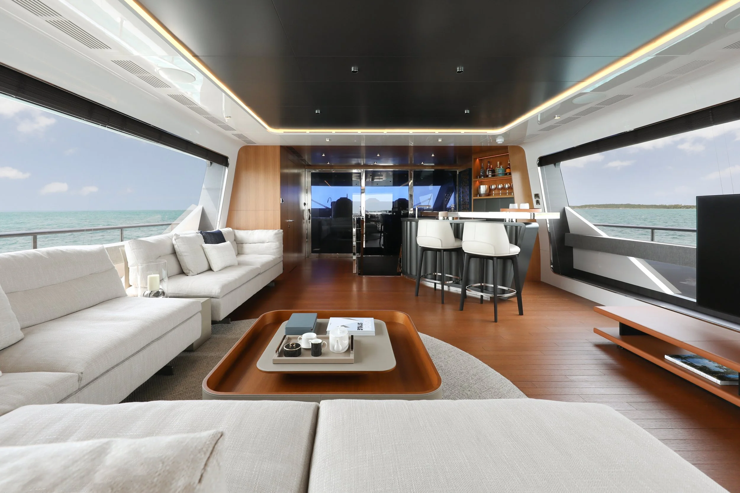 Luxury yacht interior with a large beige sofa, wooden coffee table, and a bar area with white bar stools, overlooking the ocean through wide windows.