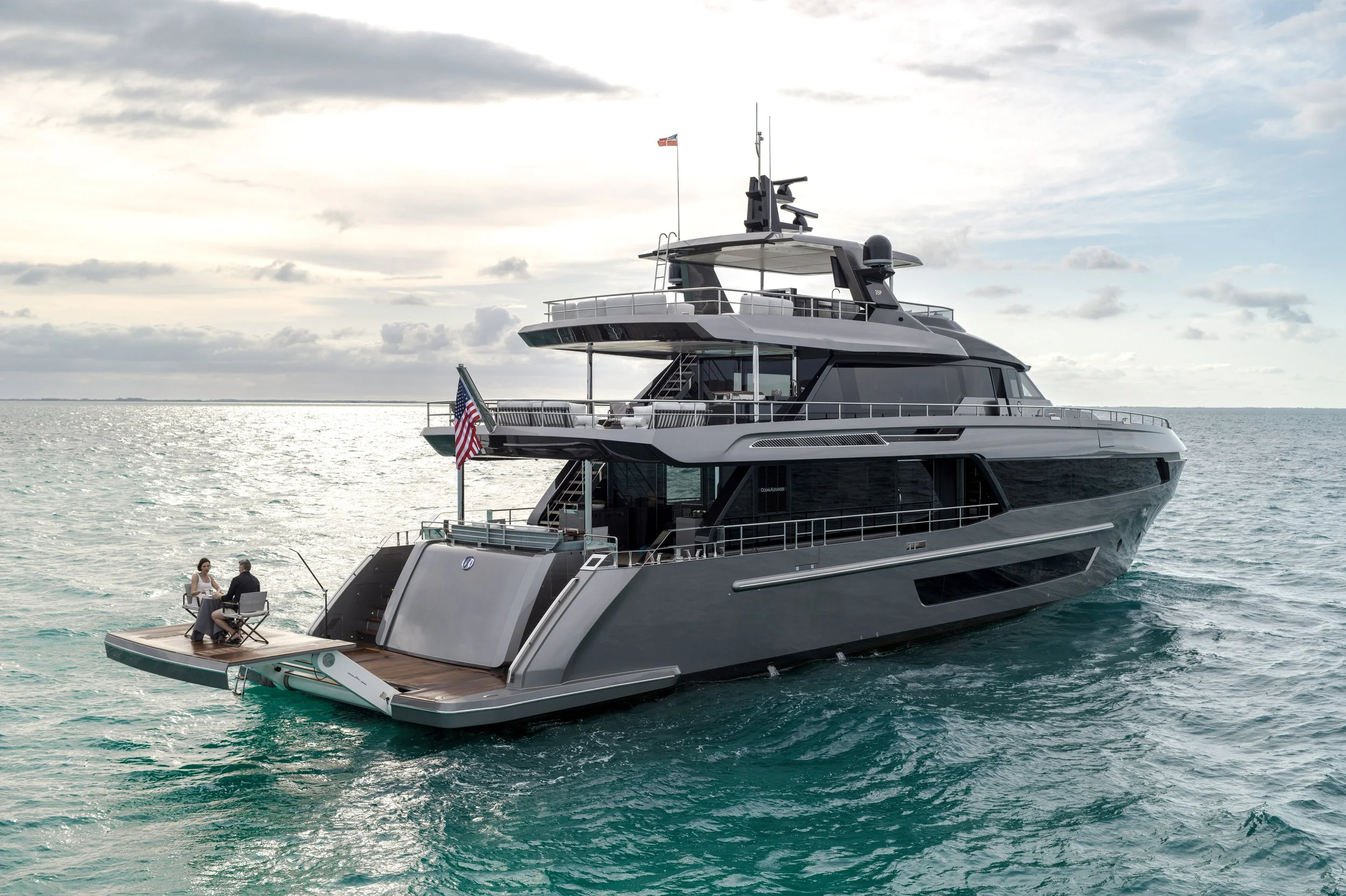Large luxury yacht on the ocean with two people seated on the deck at the stern, under a cloudy sky.