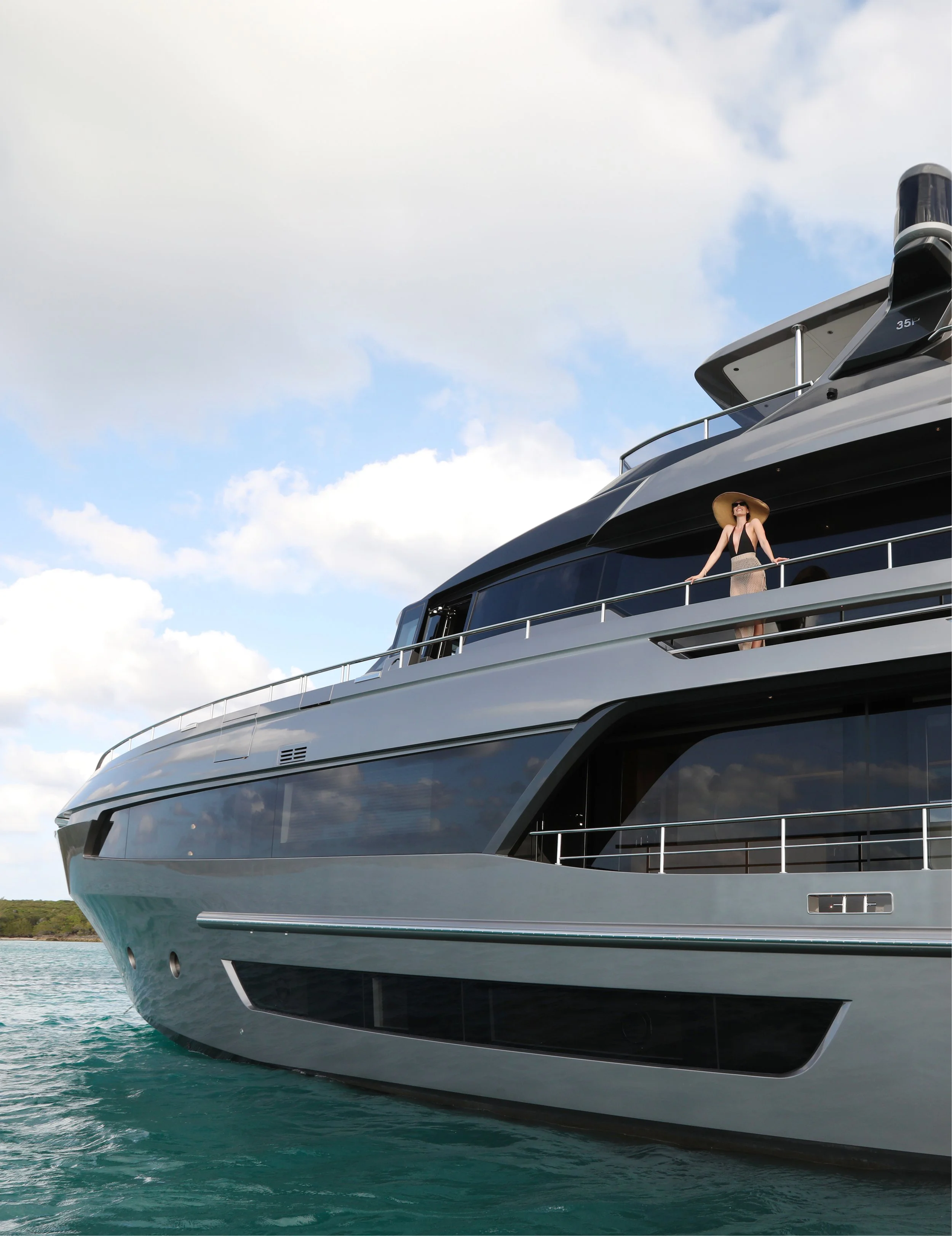 A woman in a sunhat and sunglasses standing on the deck of a large, modern yacht, overlooking the water with a cloudy sky in the background.