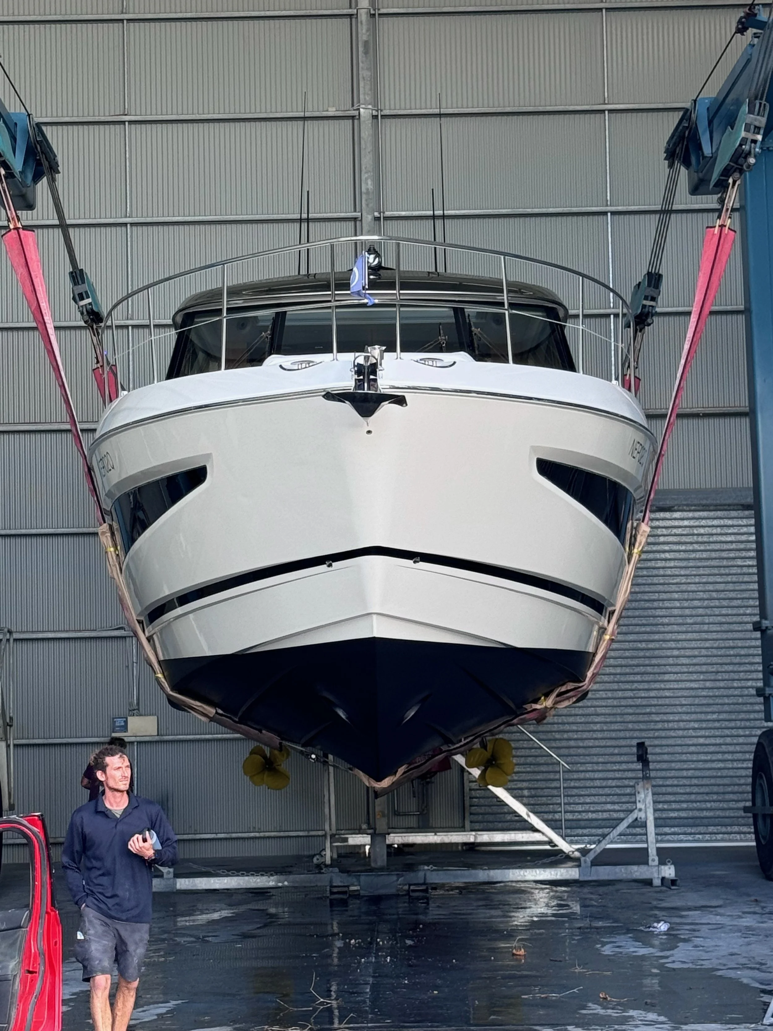 A large white yacht is inside a boat storage or maintenance facility, supported by a boat lift. A man wearing a navy blue shirt and shorts is walking nearby, holding a phone.