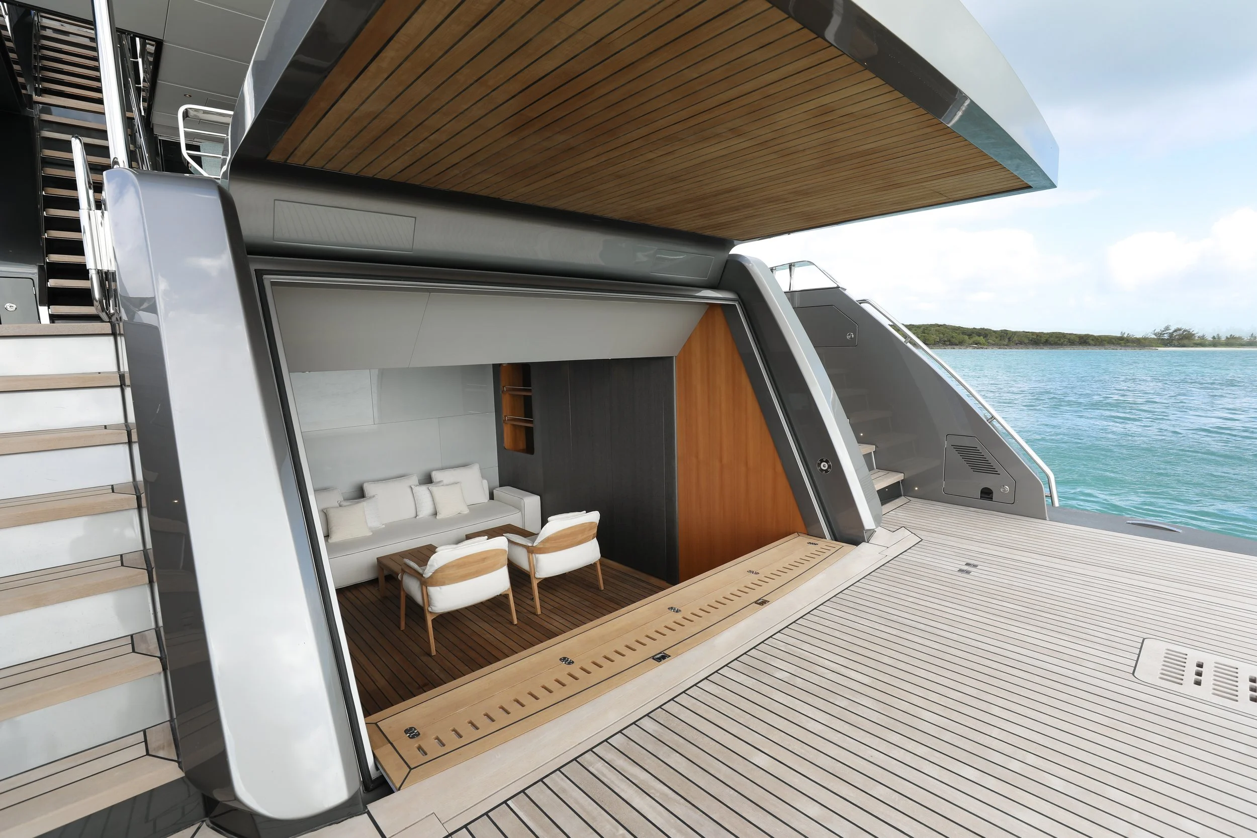 Luxury yacht with open sliding door revealing a modern outdoor lounge area with white sofa and chairs, wooden flooring, overlooking water and sky.