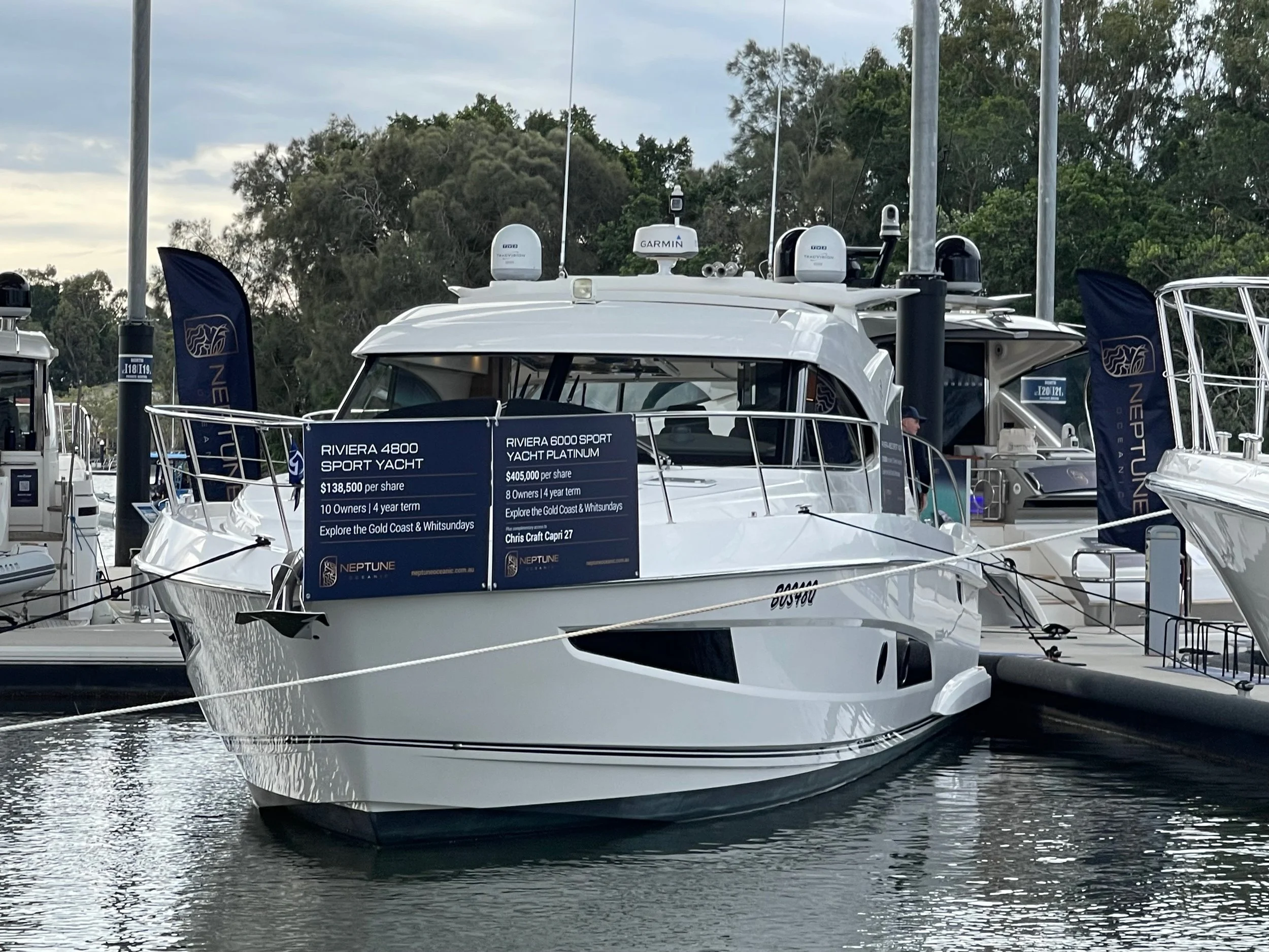 A yacht docked at a marina with informational signs and flags, surrounded by other boats and trees in the background.