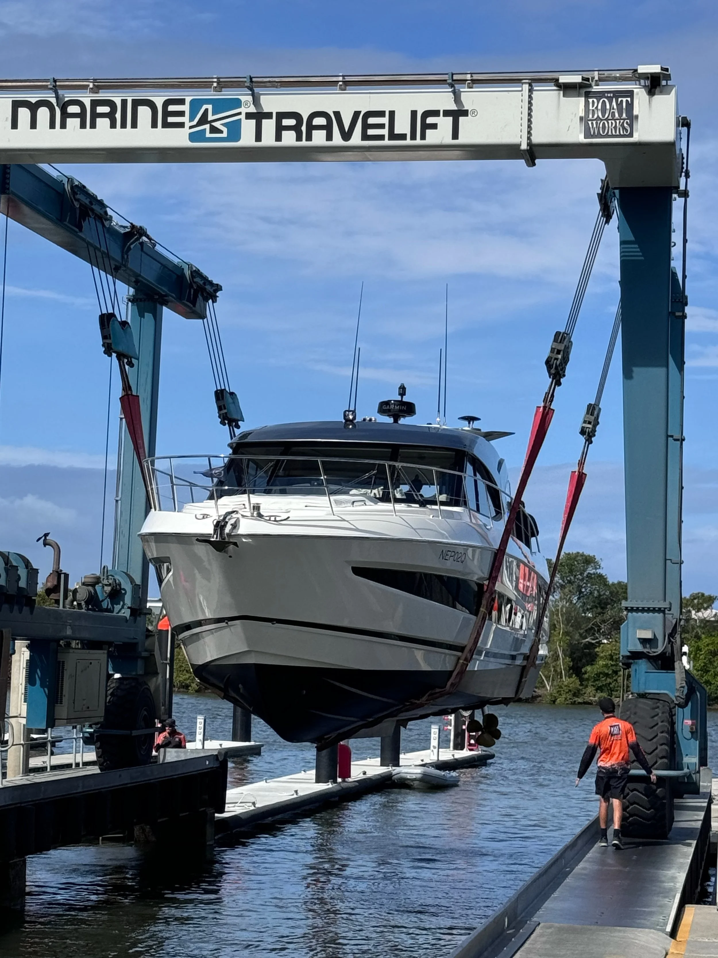 A large boat is being lifted out of the water by a Marine Travelift boat hoist on a dock. The sky is clear and blue, with some clouds, and there are trees in the background.
