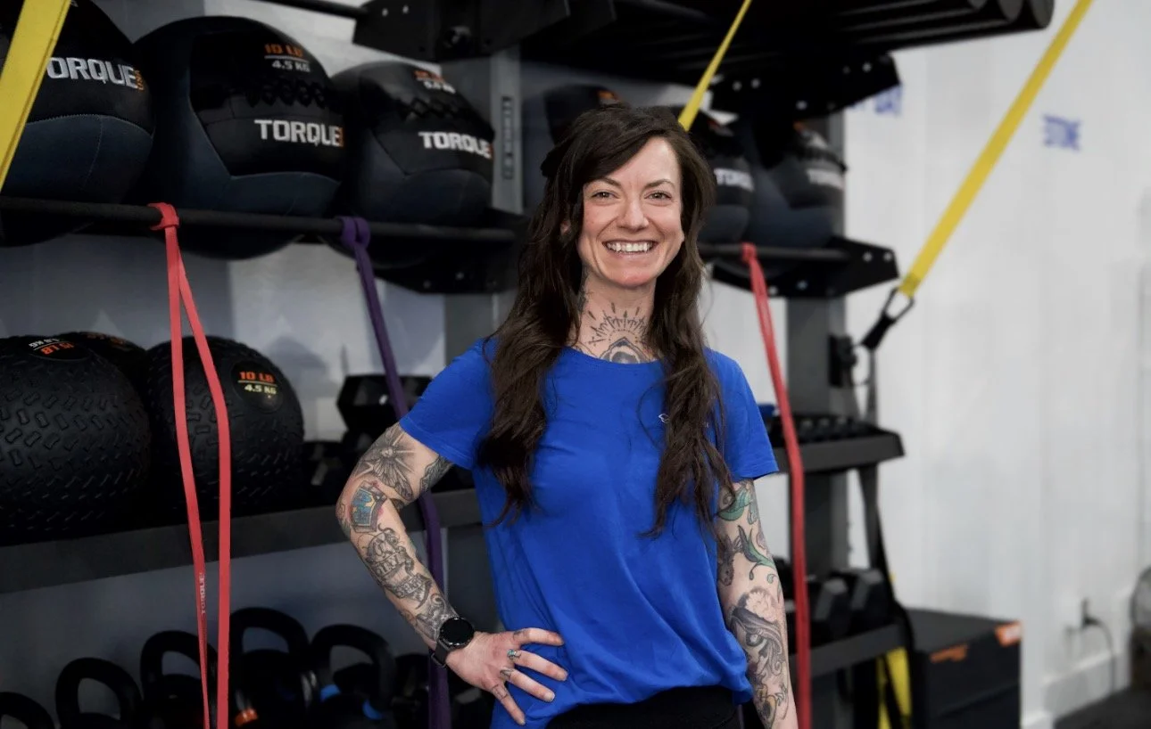 A woman with a big bun hairstyle, tattoos on her arms and neck, wearing a blue T-shirt and smiling.