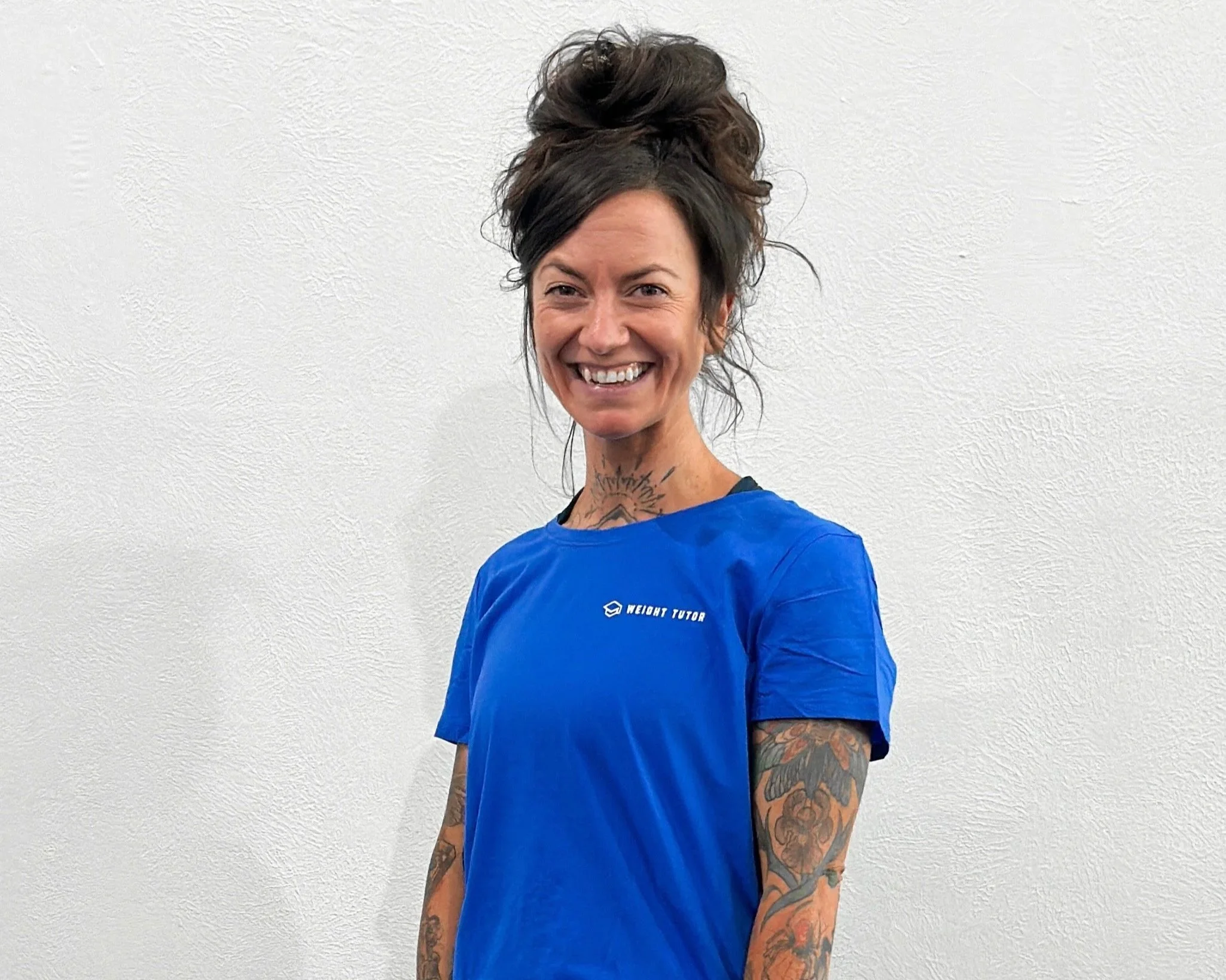 A woman with a big bun hairstyle, tattoos on her arms and neck, wearing a blue T-shirt and smiling.