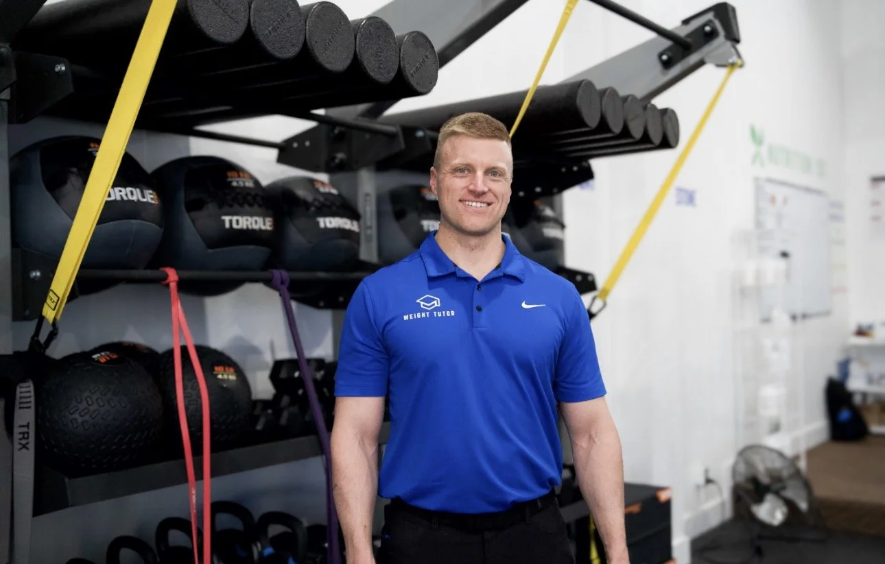 A man smiling in a gym wearing a blue shirt with 'Weight Tutor' logo and a gray and blue cap.