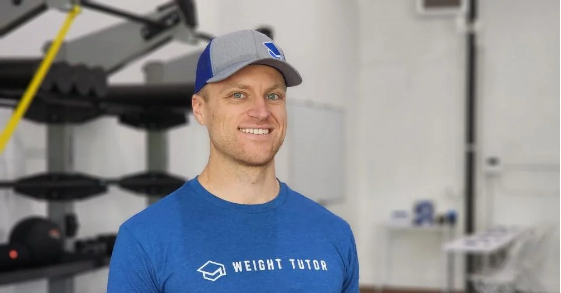 A man smiling in a gym wearing a blue shirt with 'Weight Tutor' logo and a gray and blue cap.