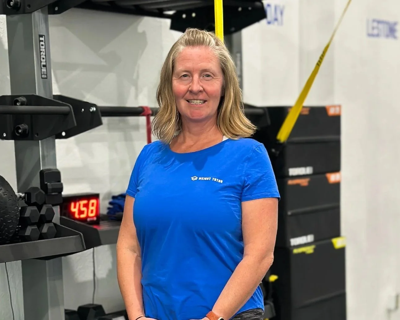 A smiling woman with shoulder-length blonde hair wearing a blue workout shirt standing in a gym near workout equipment.