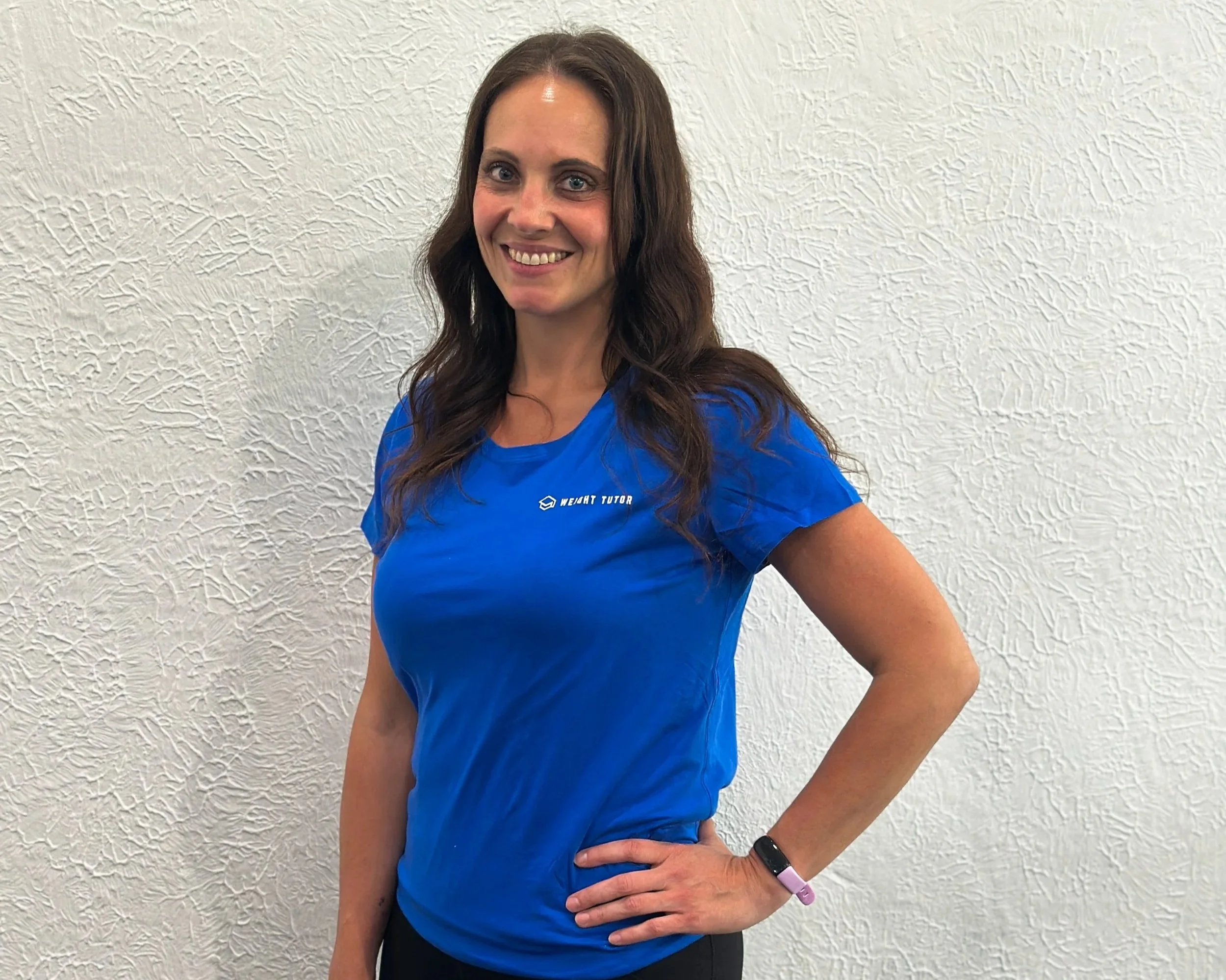 A woman with long brown hair smiling, wearing a blue t-shirt and a fitness tracker, standing with her hand on her hip against a textured white wall.