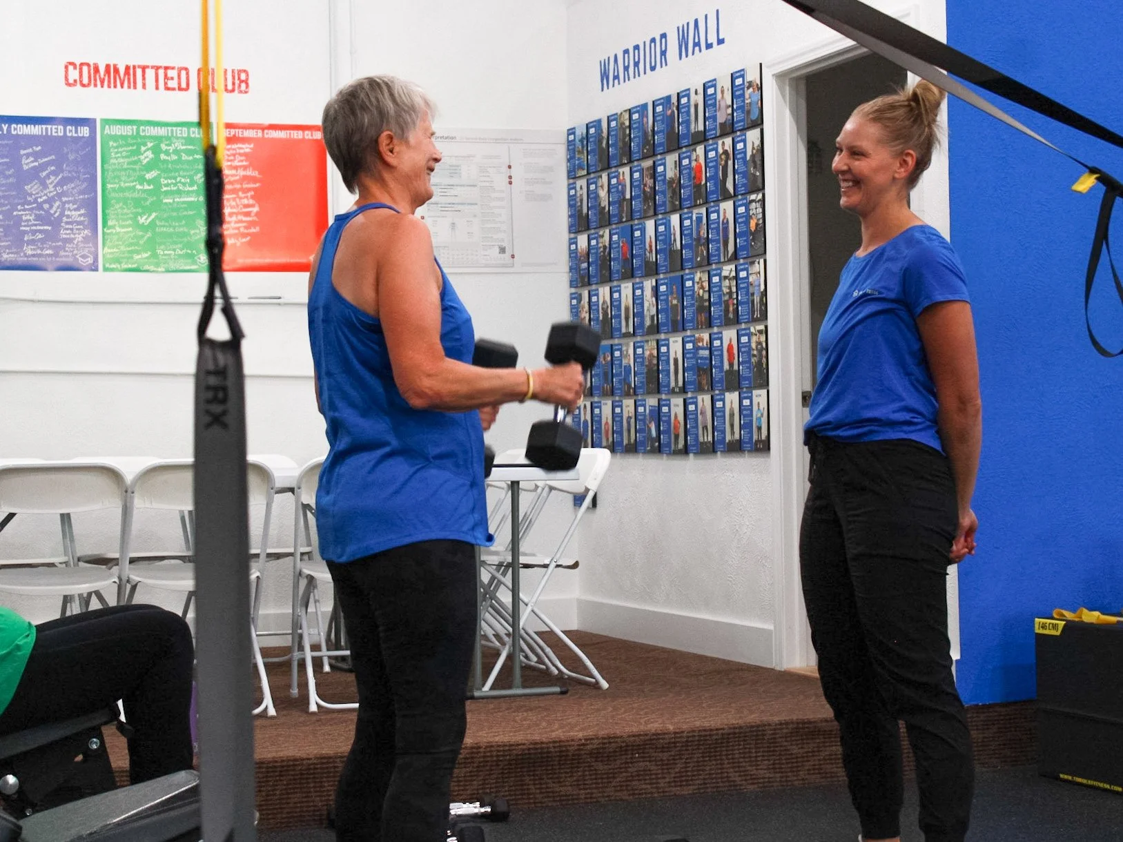 Two women in blue workout shirts smiling and talking in a gym; one is holding a dumbbell, and the wall behind features a fitness award or achievement display.