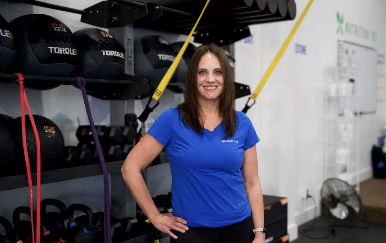 A woman with long brown hair smiling, wearing a blue t-shirt and a fitness tracker, standing with her hand on her hip against a textured white wall.