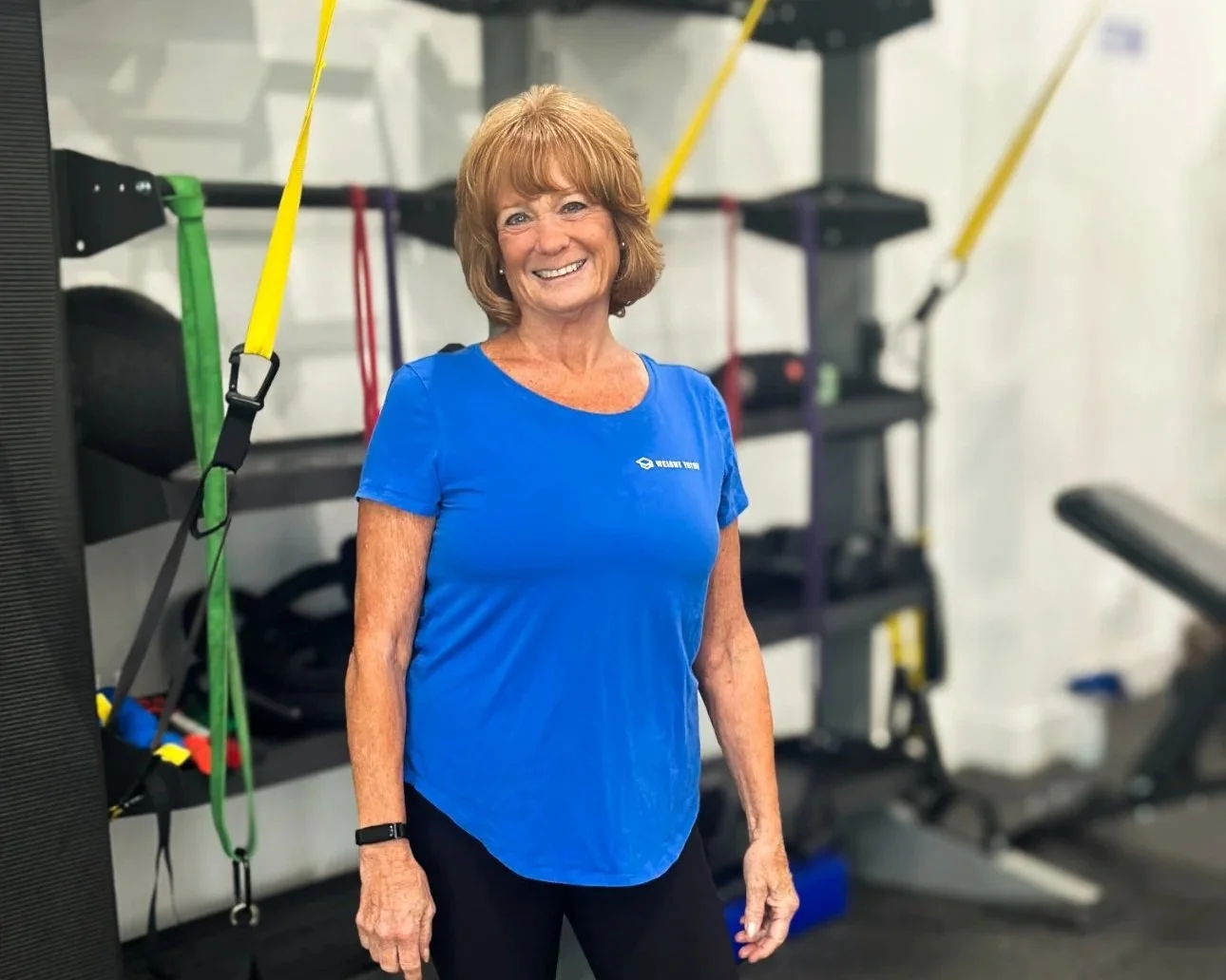 A smiling woman with short, reddish hair in workout attire standing in a gym near exercise equipment with resistance bands and weight racks.