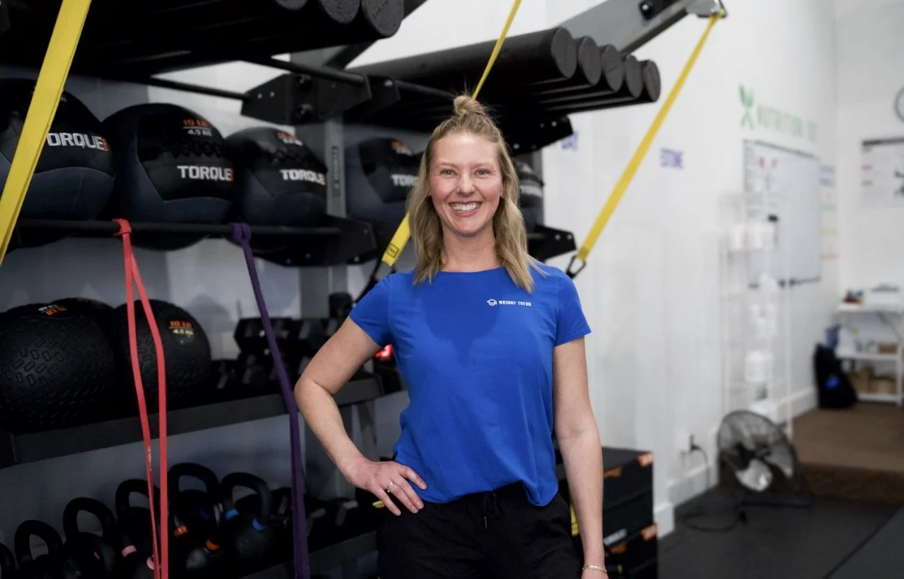 A woman smiling in a gym, wearing a blue shirt with a logo that reads 'Weight Tutor', standing next to gym equipment.