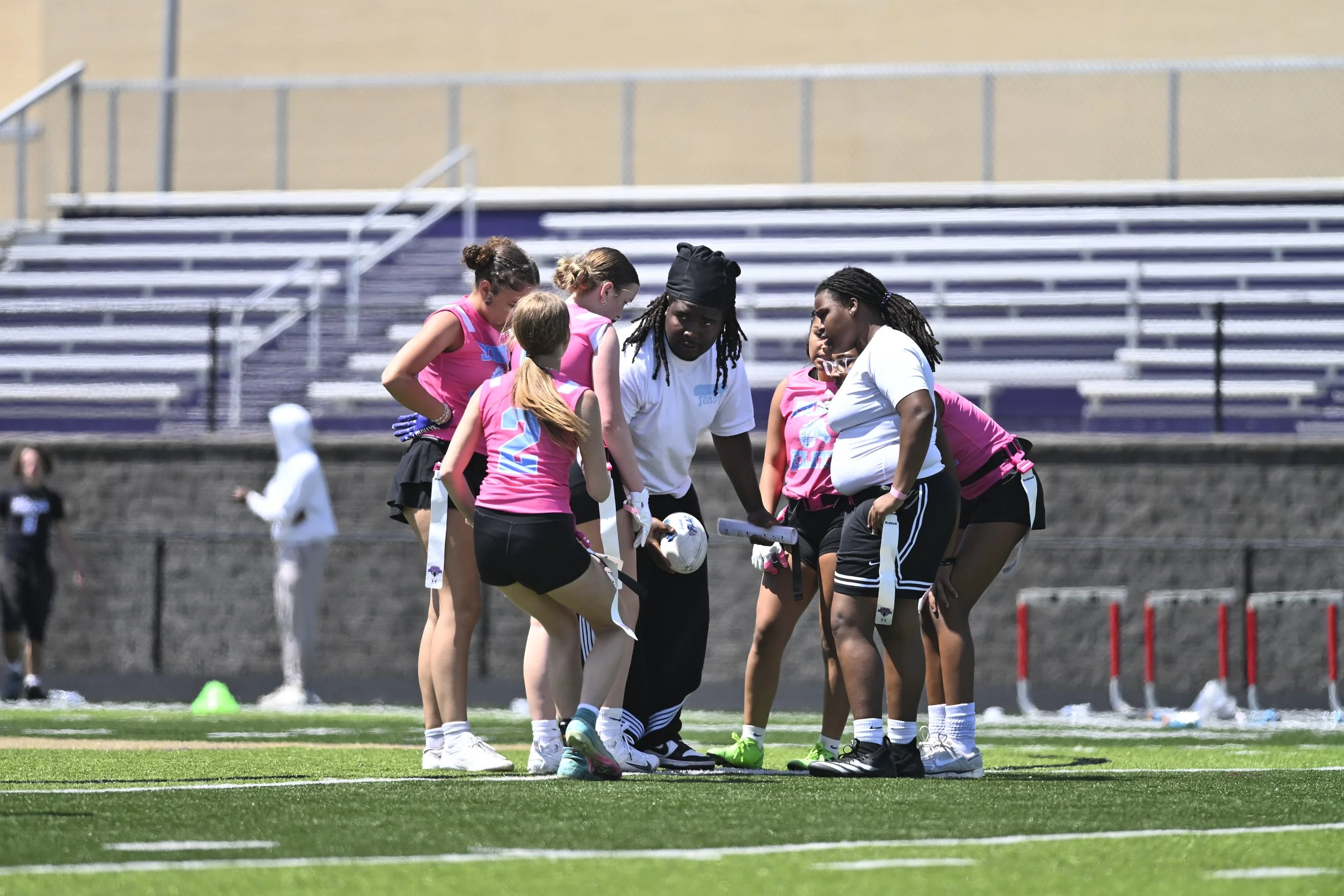 A group of female football players, dressed in pink and black, are gathered around their coach on the football field during a practice or game. The coach appears to be giving instructions. The background shows empty stadium seats and a few other players on the field.
