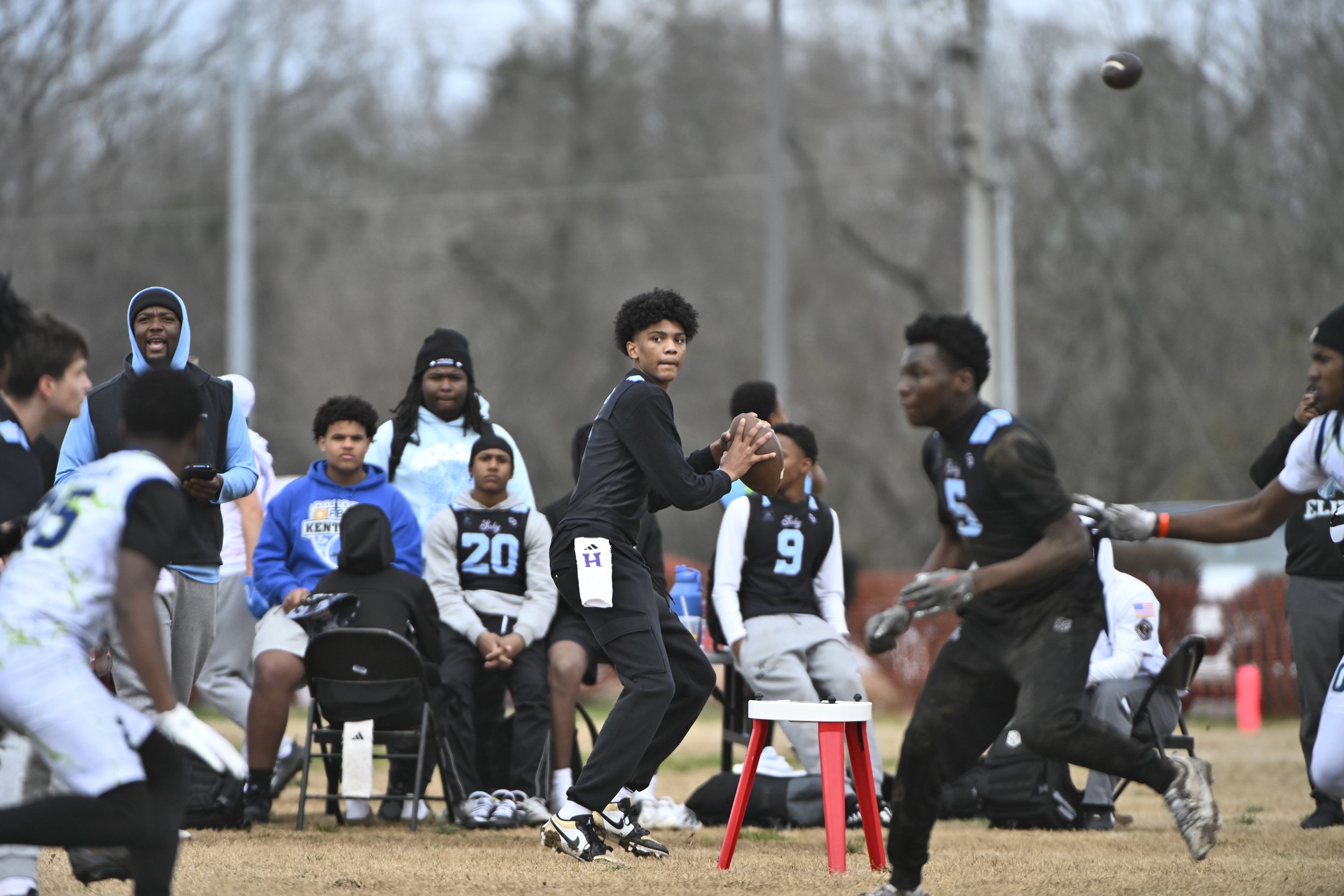 A group of young football players, some sitting and some standing, watching or participating in a game outdoors on a cloudy day. One player is holding a football and standing near a stool, with trees and utility poles in the background.